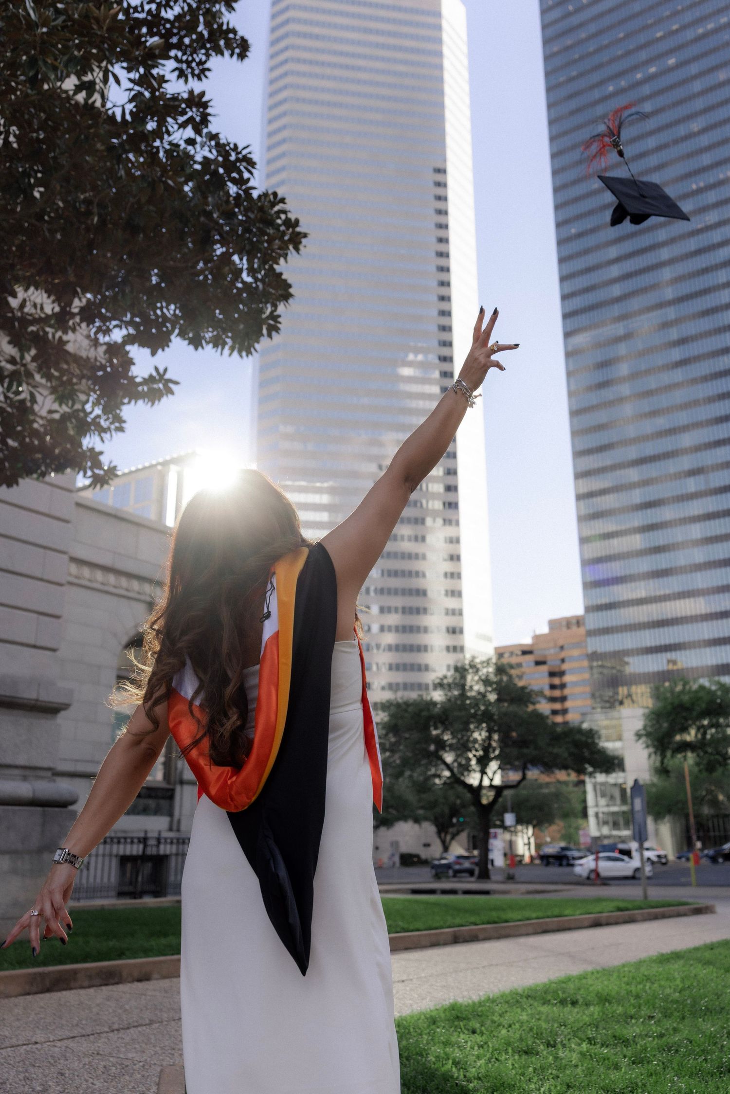 Graduate in black robe tossing cap into air near glass skyscrapers with sunlight flare.