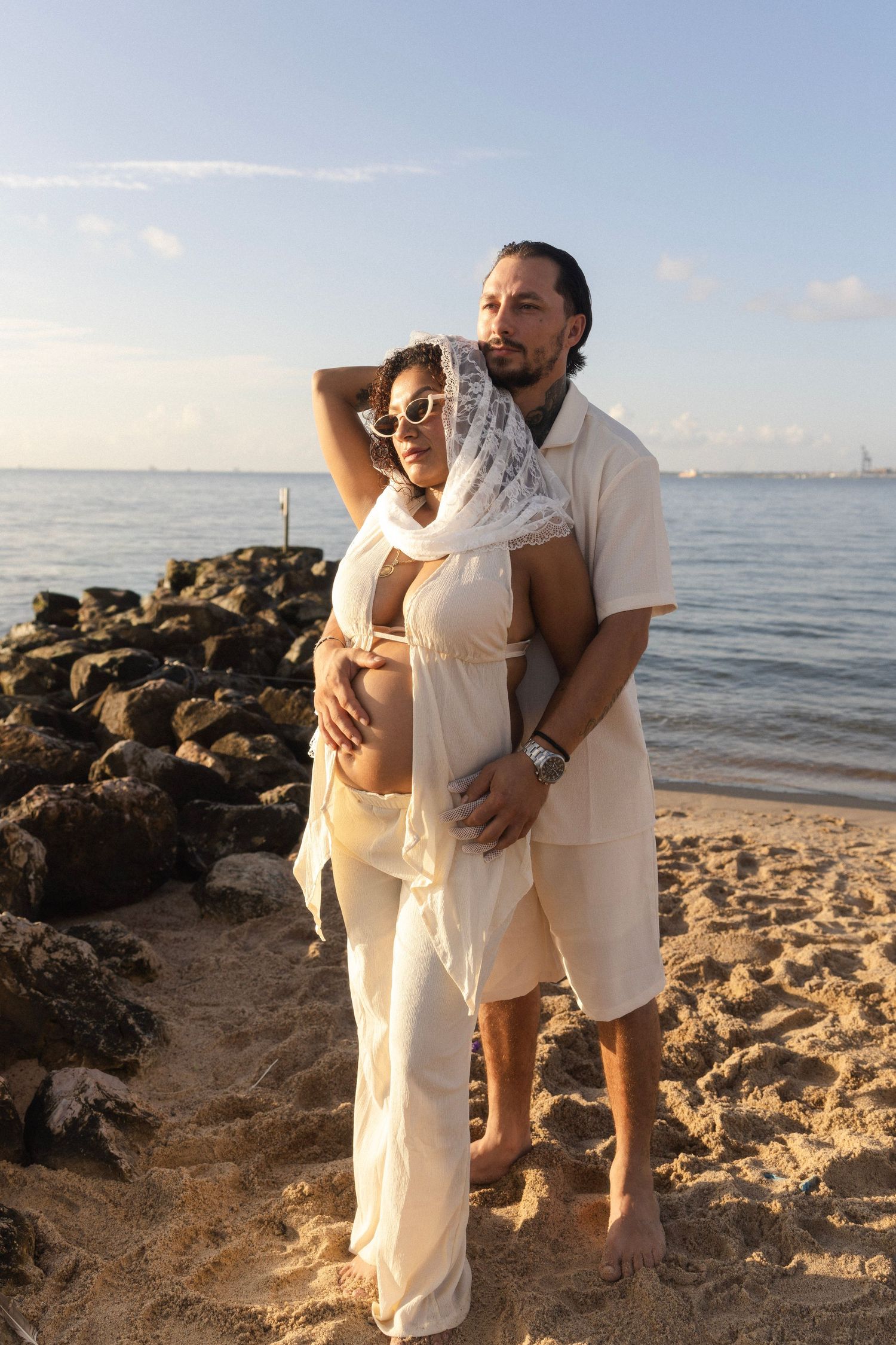 Pregnant couple poses on sandy beach near rocky shoreline, woman in white outfit with lace headscarf, man embracing her bump.