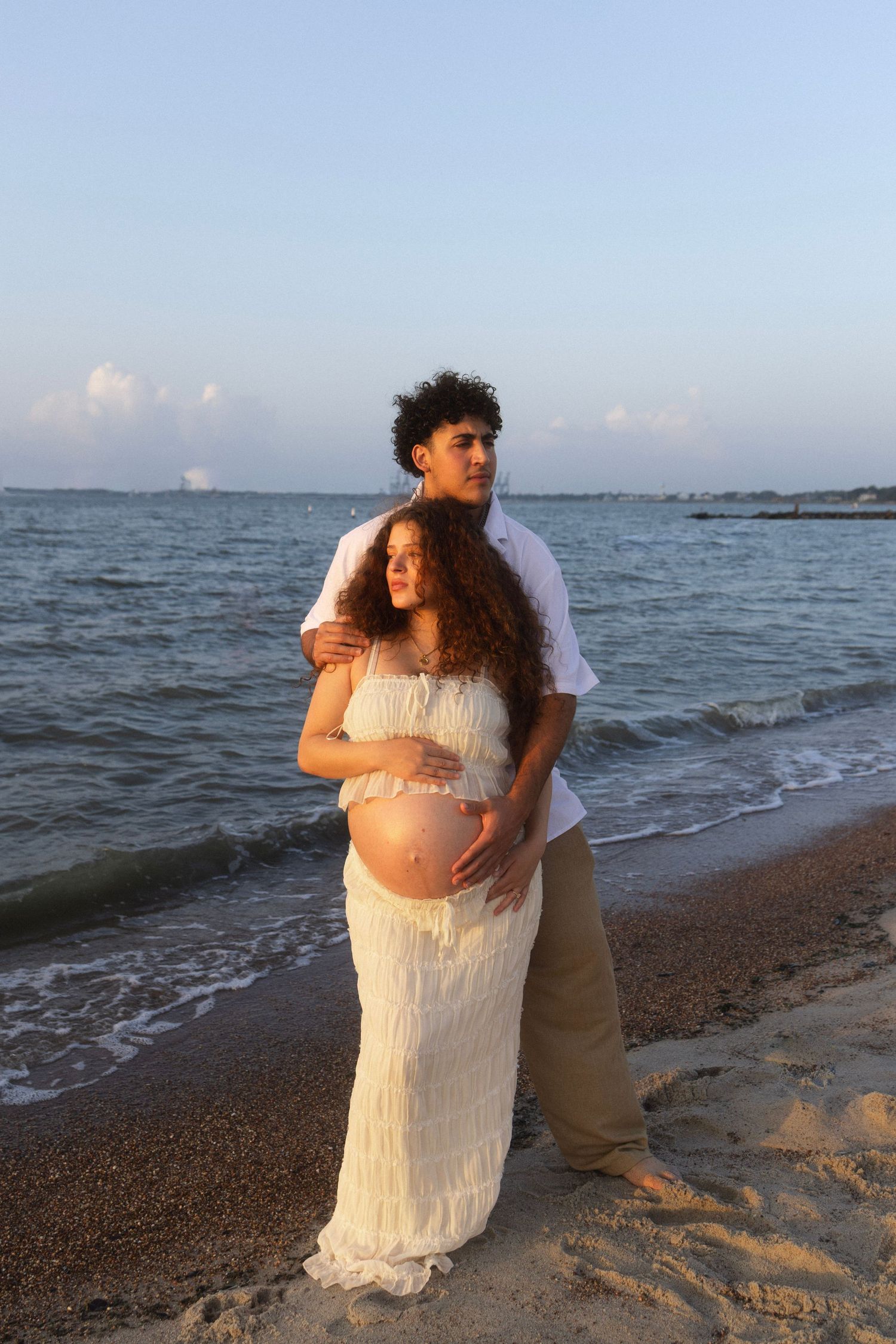 Pregnant couple embracing on beach at sunset, woman in white fringe dress cradling baby bump, man in white shirt behind her.