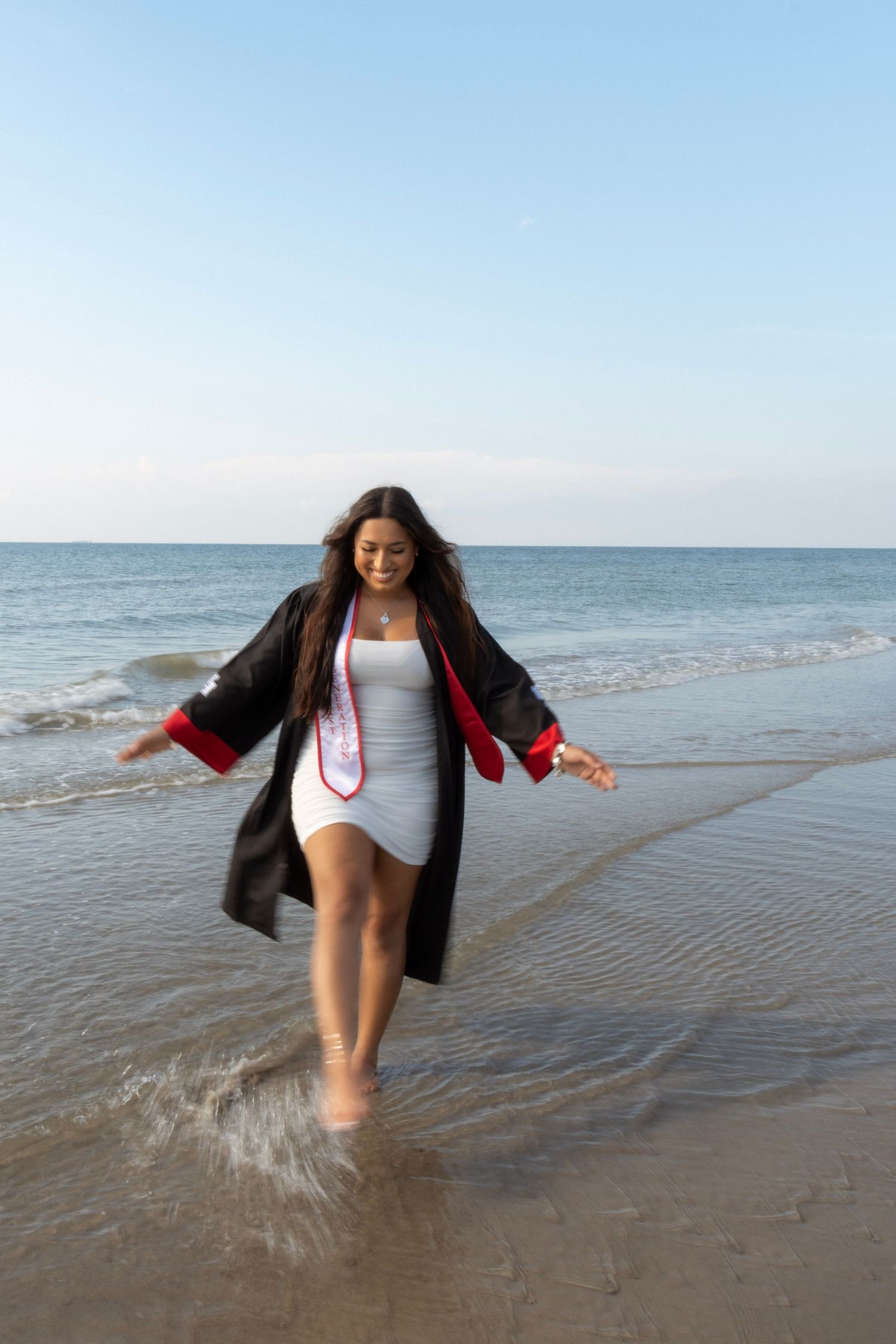 Female graduate in cap and gown walking joyfully through ocean waves on sandy beach.