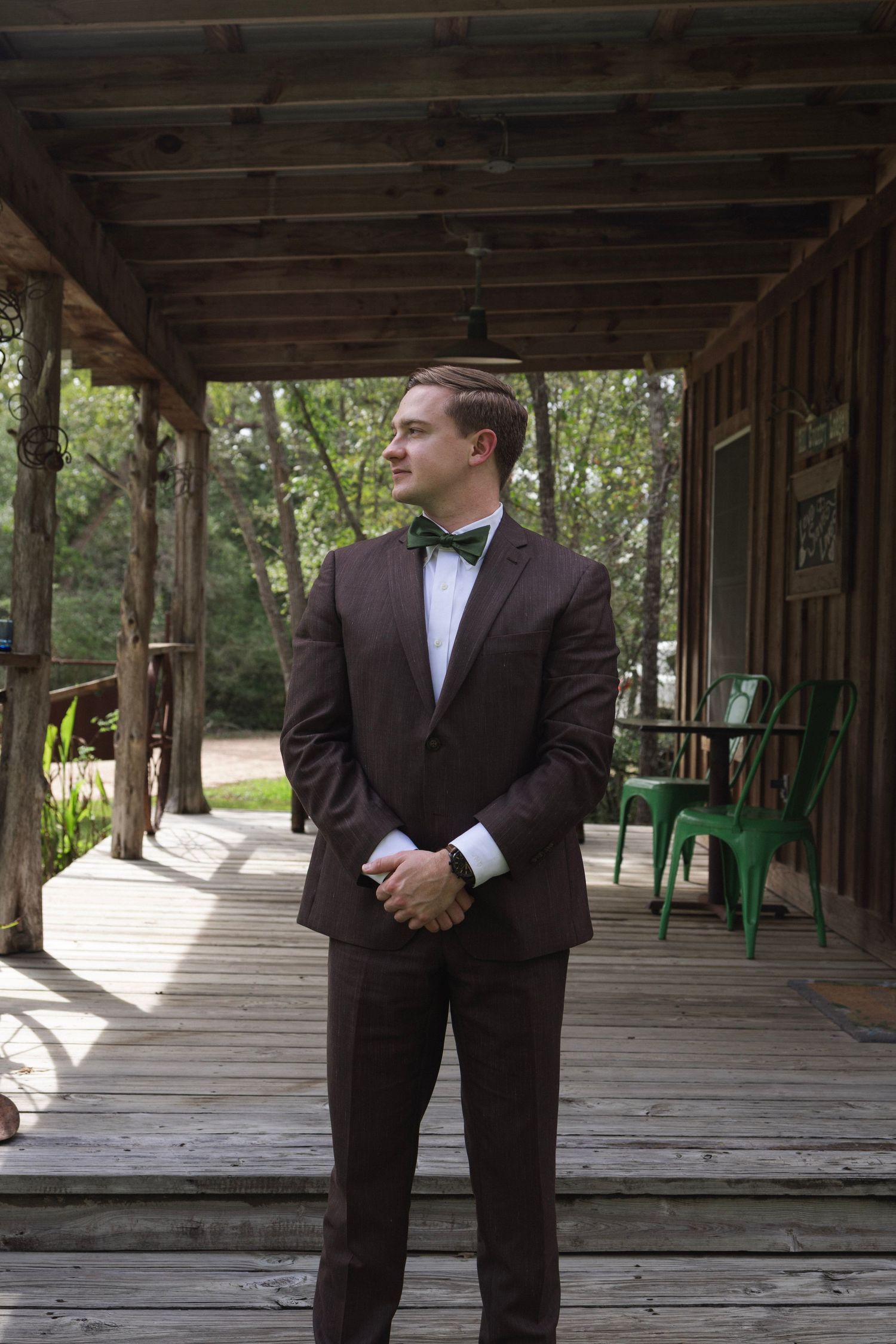A groom in a brown tuxedo with green bow tie stands on a rustic wooden porch surrounded by trees.