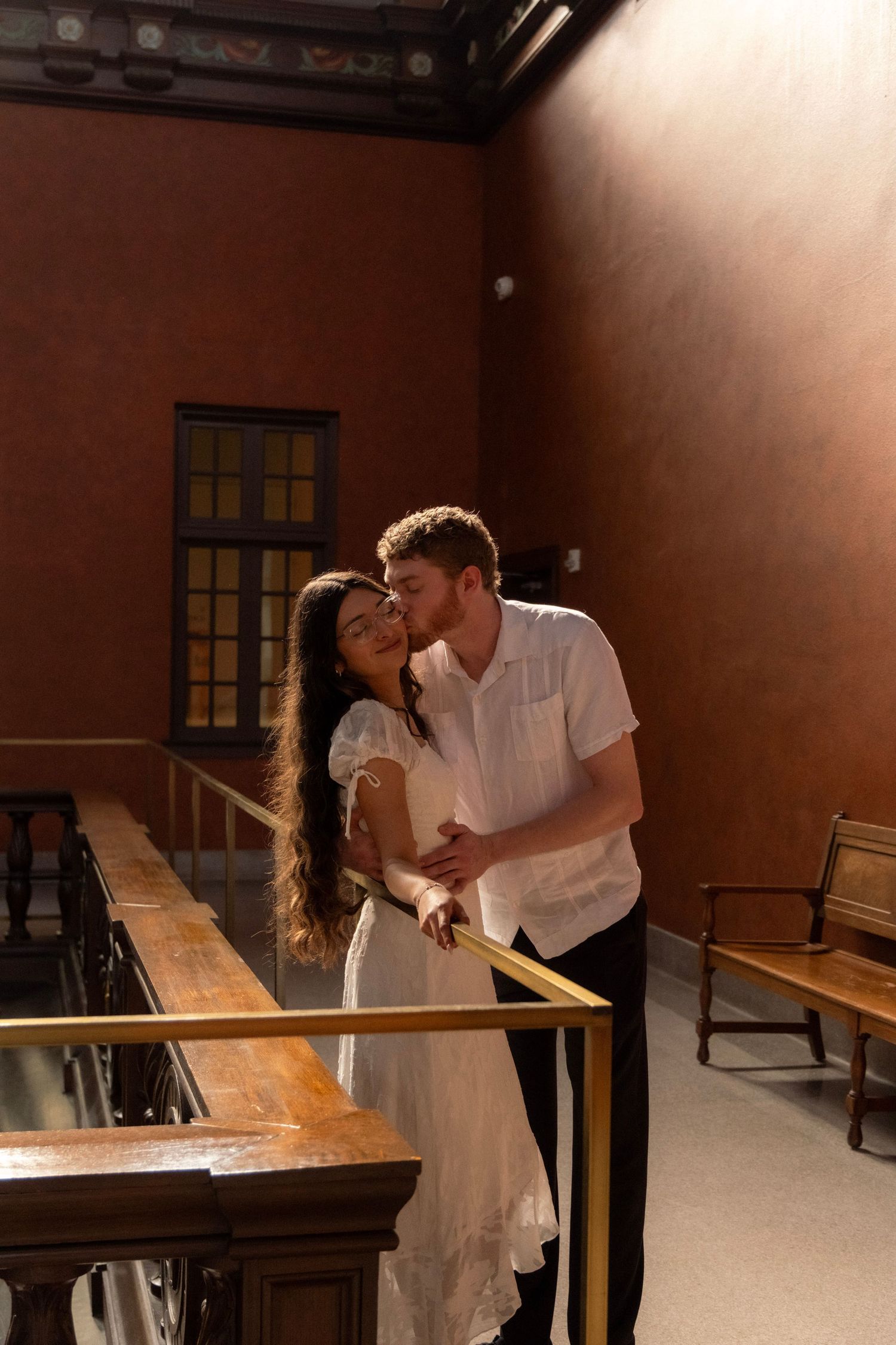 Couple sharing intimate moment on golden railing balcony inside warmly lit historic building.