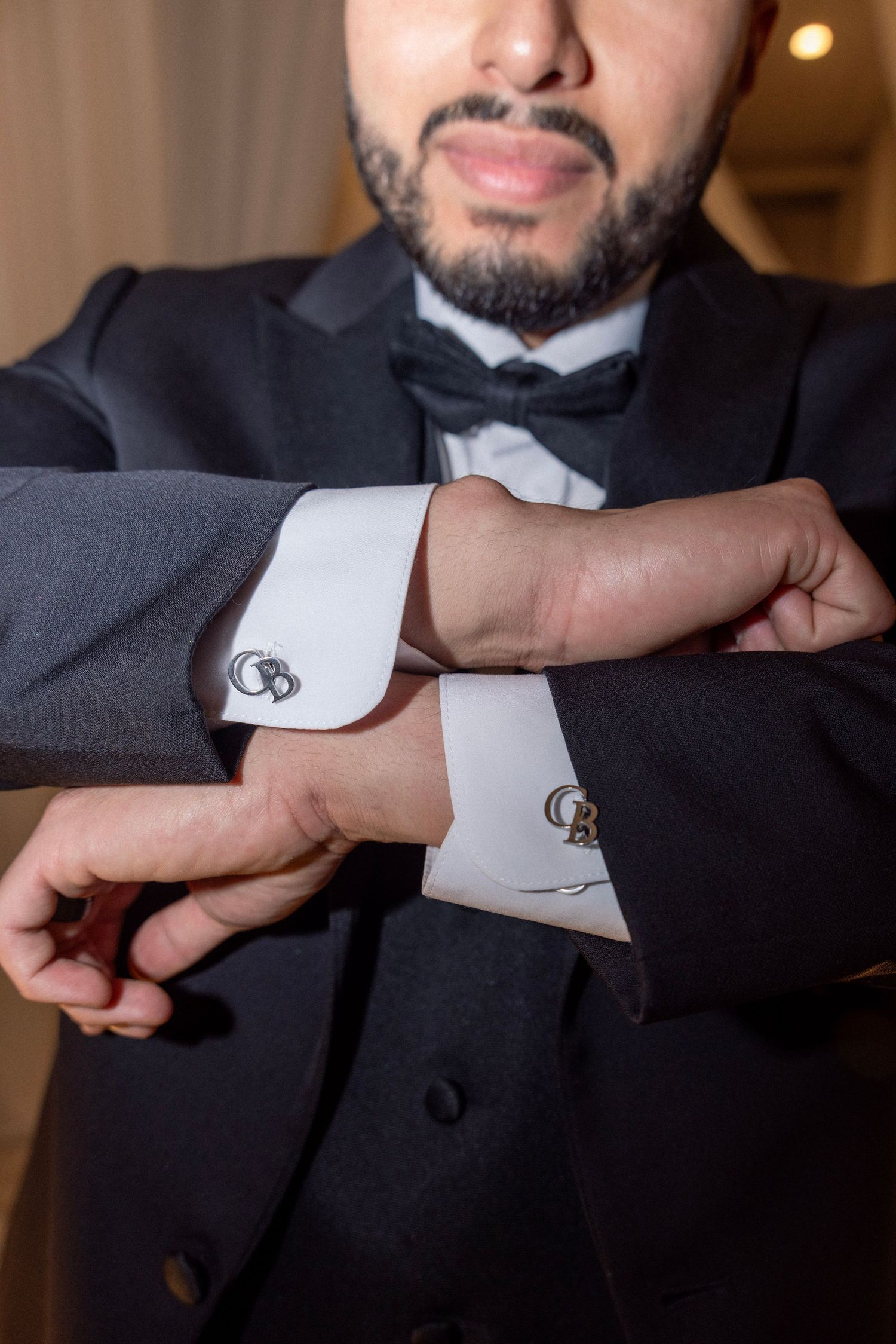 Bearded groom in tuxedo adjusting monogrammed cufflinks on white dress shirt with black bow tie.