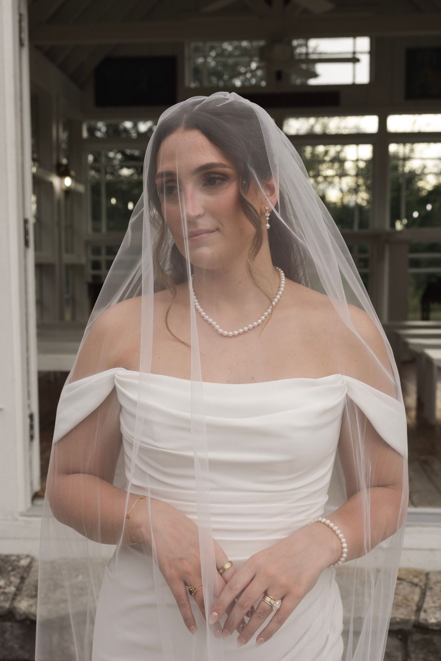 Bride in white off-shoulder gown and pearl necklace wearing a flowing veil, posing elegantly outdoors.