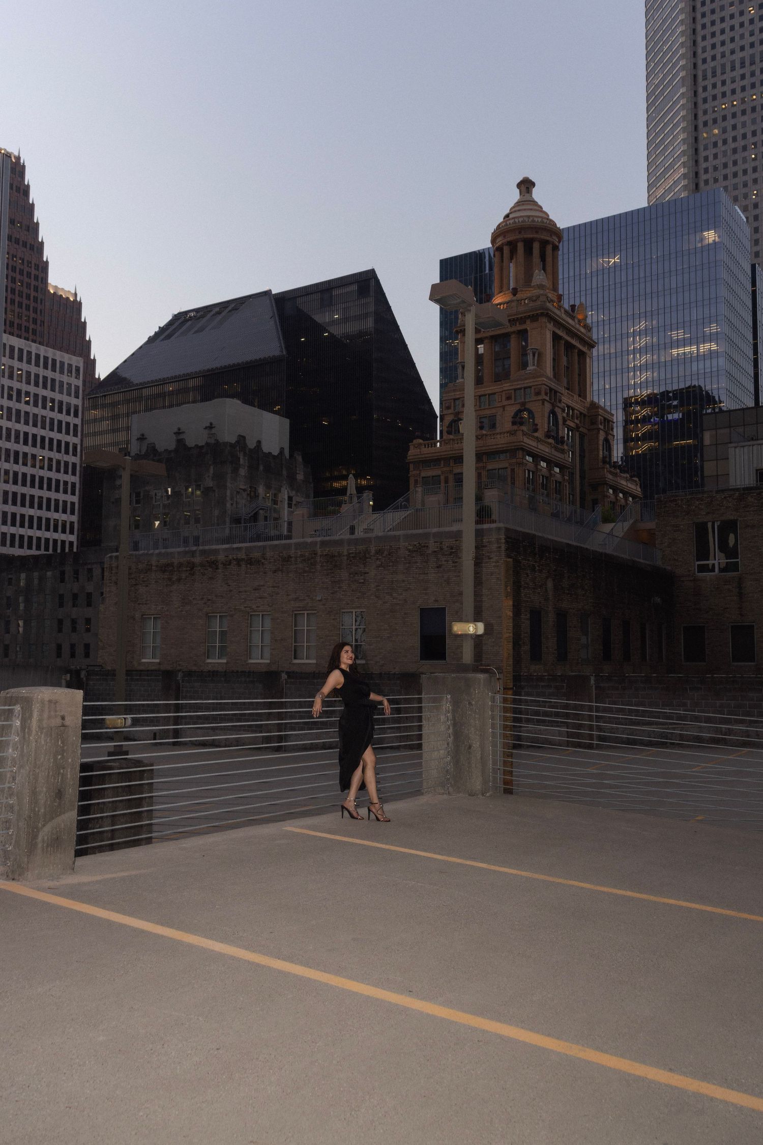 Woman in black dress posing on urban walkway surrounded by modern skyscrapers and historic brick building at dusk.