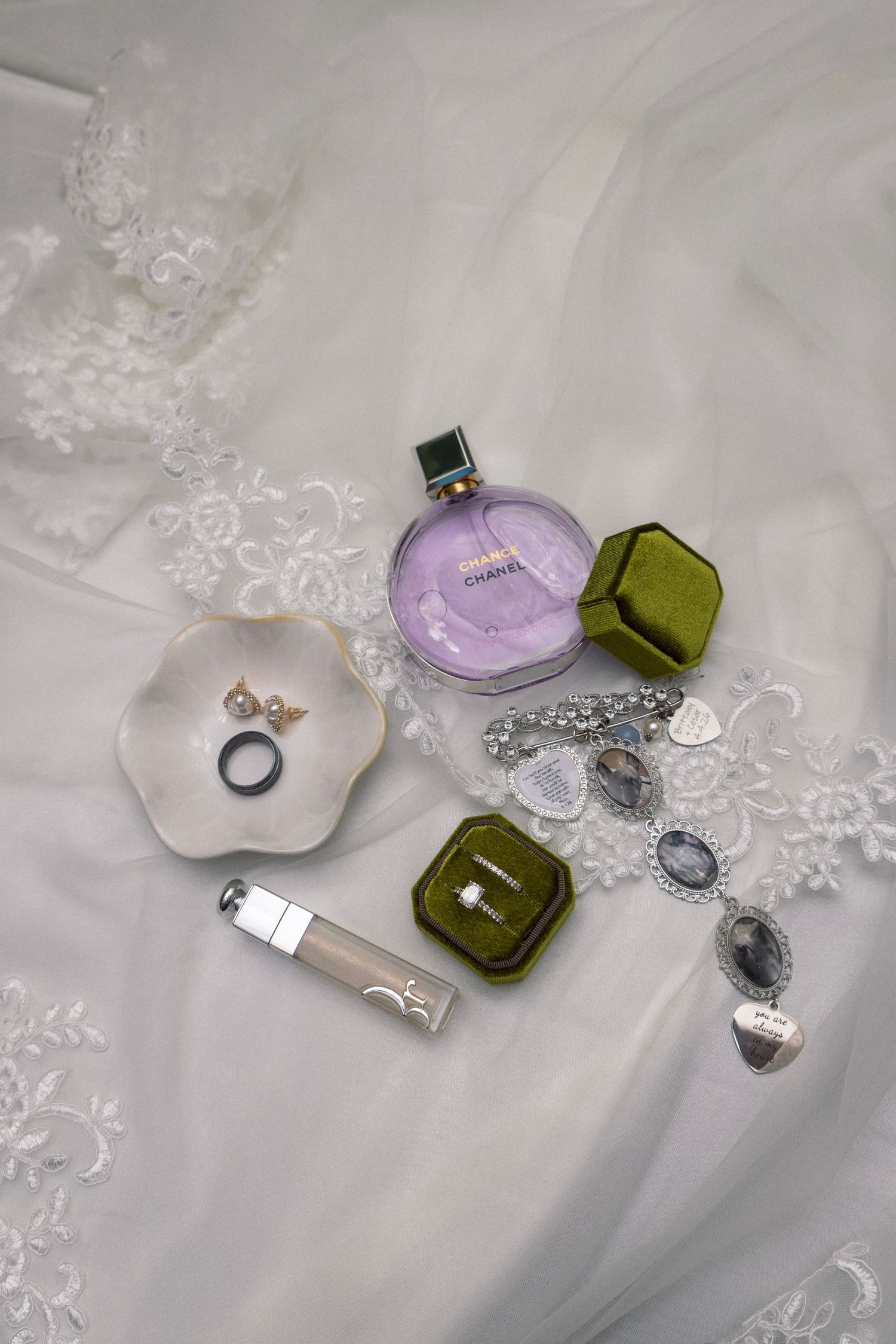 Bridal flat lay featuring Chanel perfume, Dior lip gloss, rings, and jewelry on white lace.