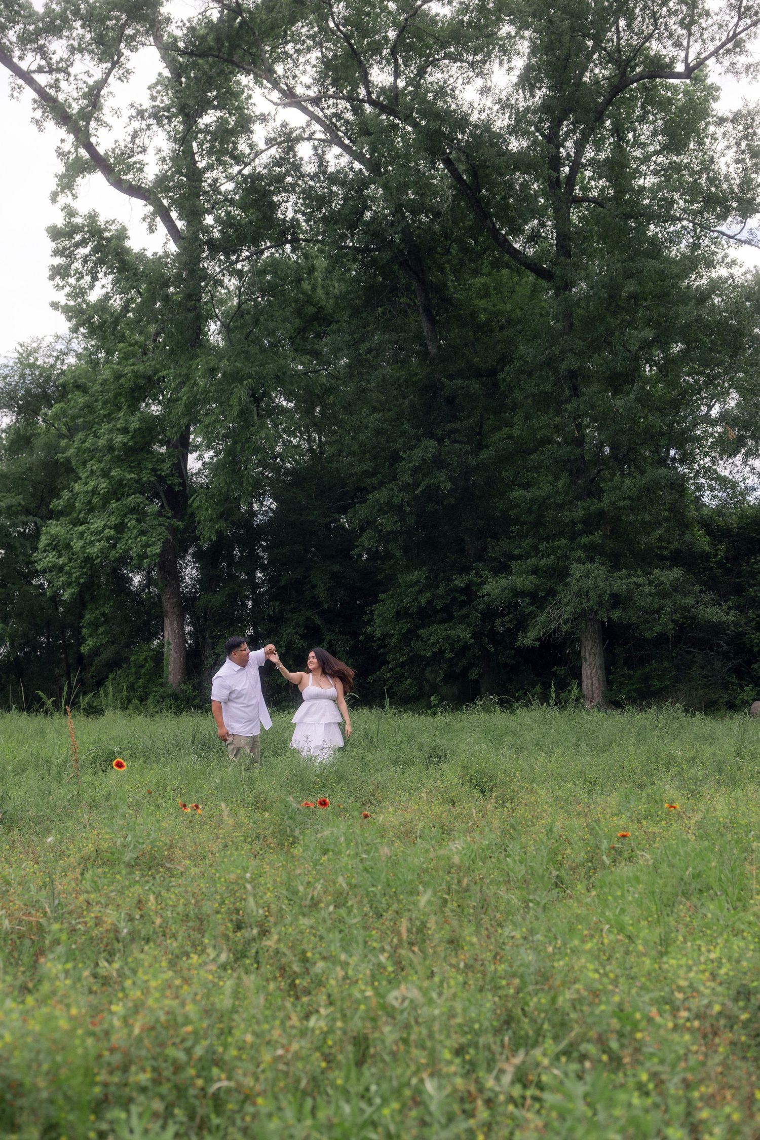 Couple dancing together in lush green meadow with wildflowers and tall trees in background.