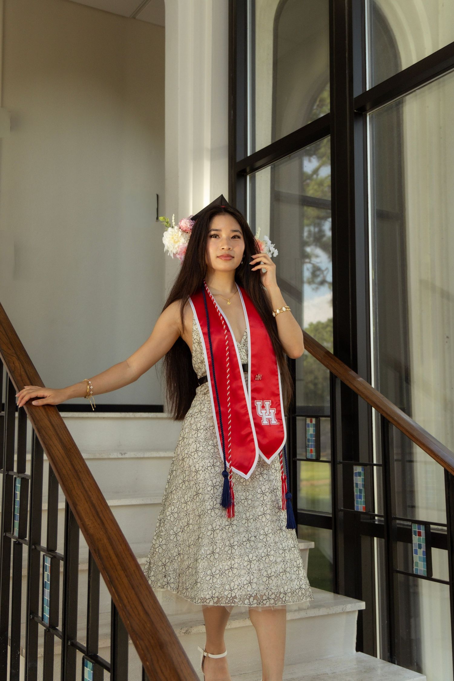 University of Houston graduate in floral dress and red stole posing on staircase by large windows.