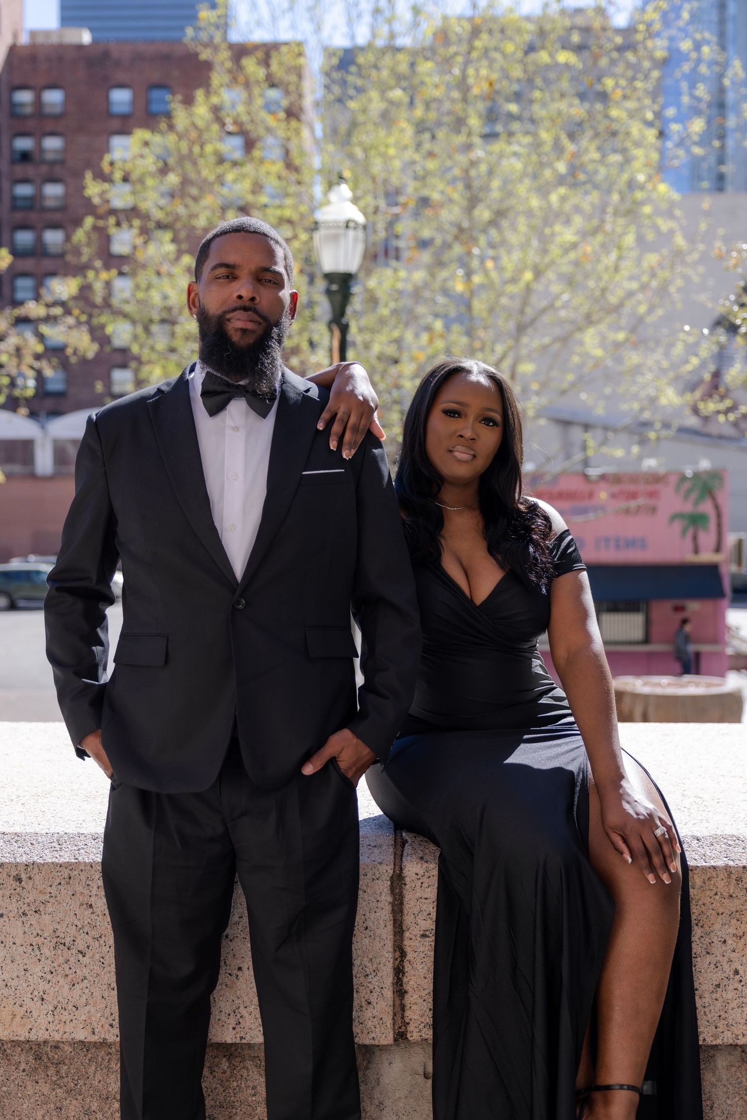 Elegant couple in black formal attire pose together in an urban outdoor setting with city buildings.