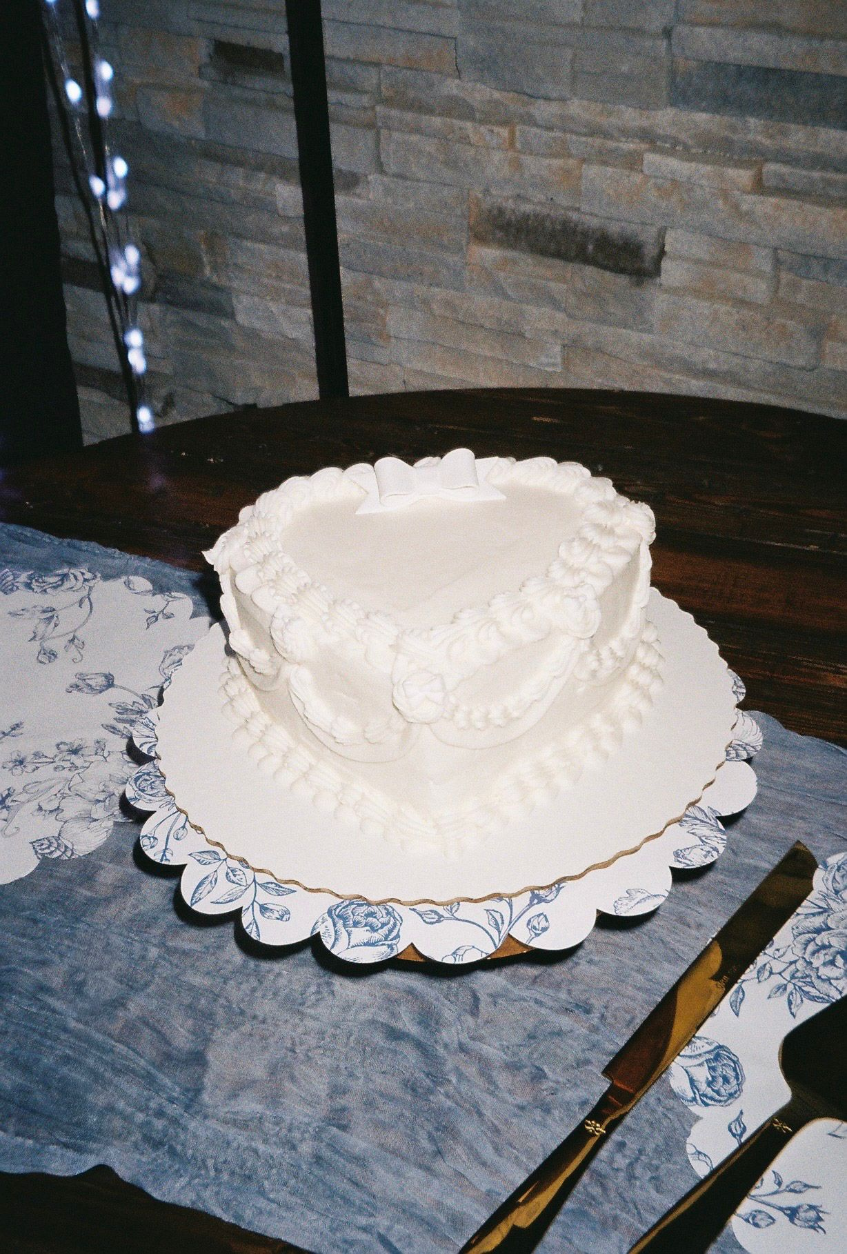 Simple elegant single-tier white wedding cake with piped floral details on a scalloped board against a stone brick wall.