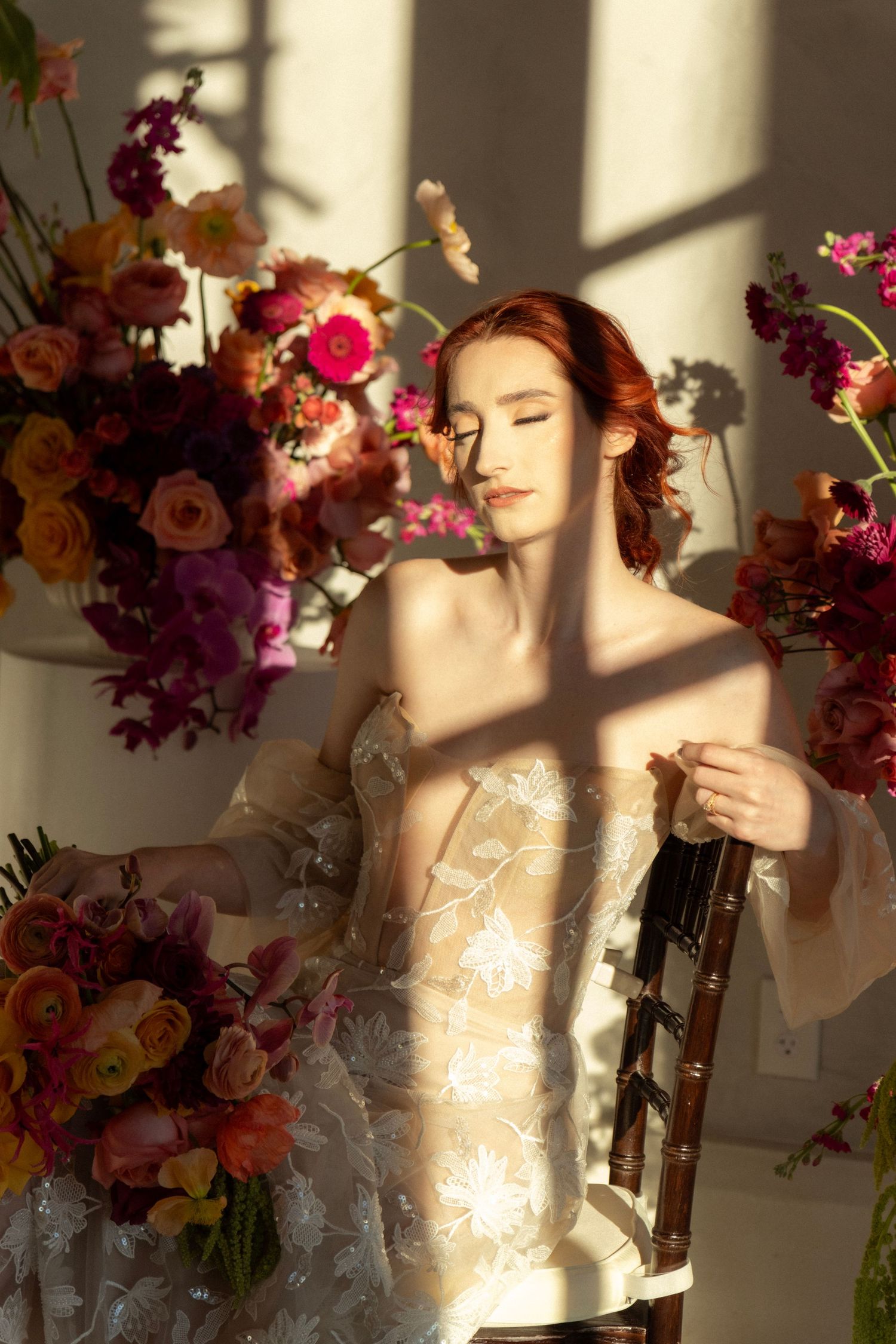 Red-haired bride in embroidered off-shoulder gown sits in sunlight surrounded by vibrant pink, orange, and purple floral arrangements.