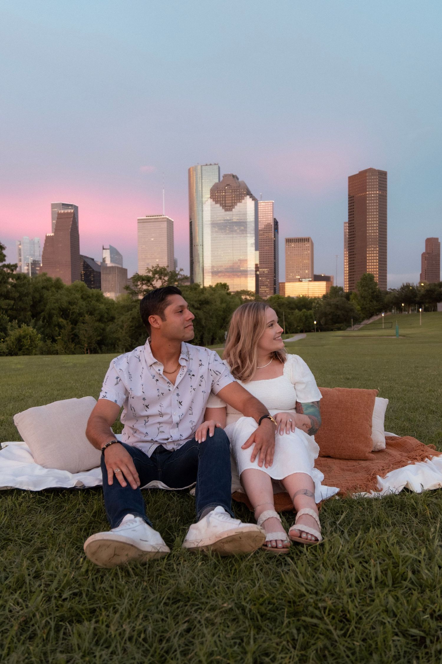 Couple sitting on a blanket in a park with the Houston skyline at sunset in the background.