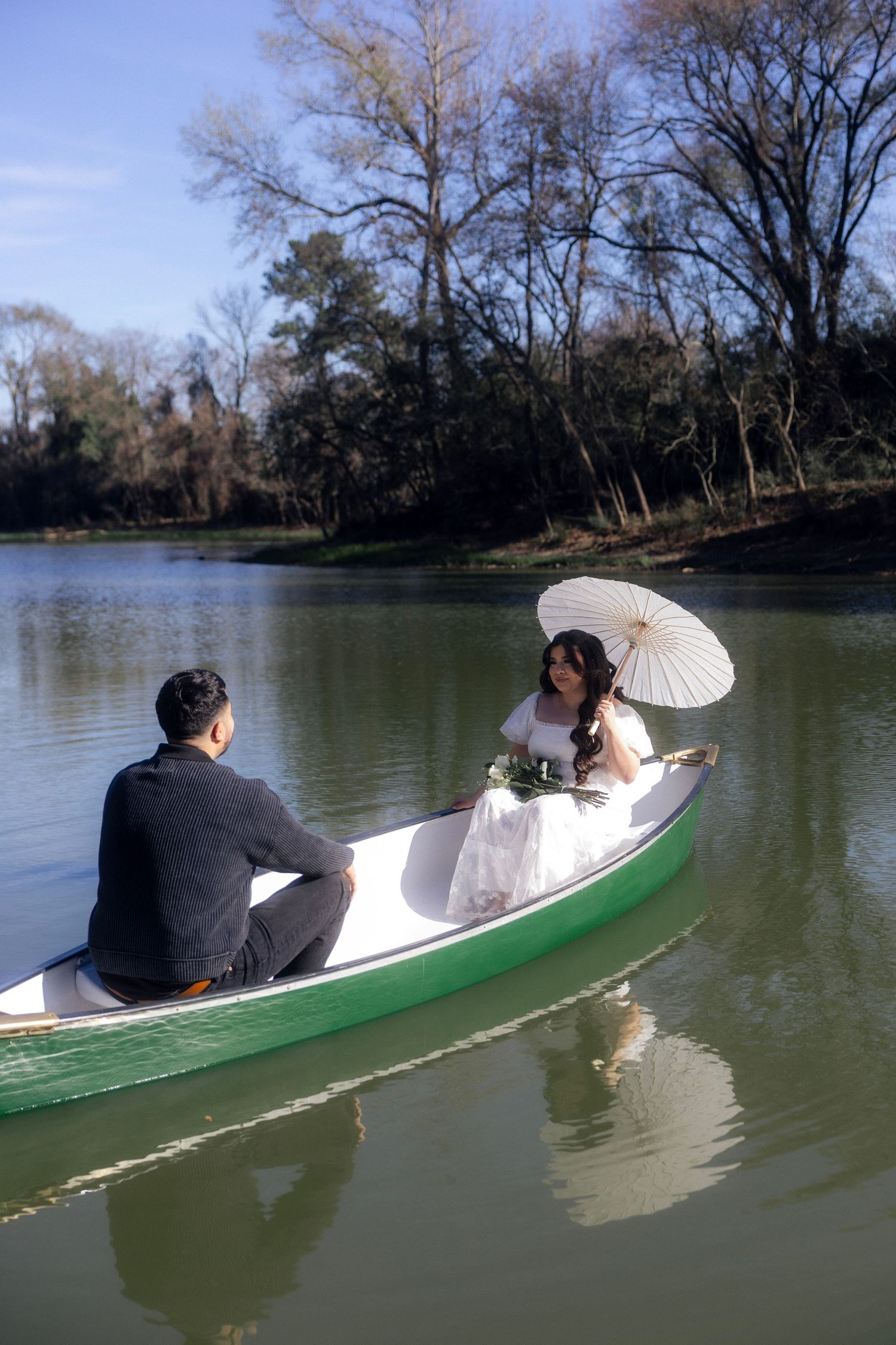 Bride with white parasol and groom in gray suit sitting in a green canoe on a calm river.