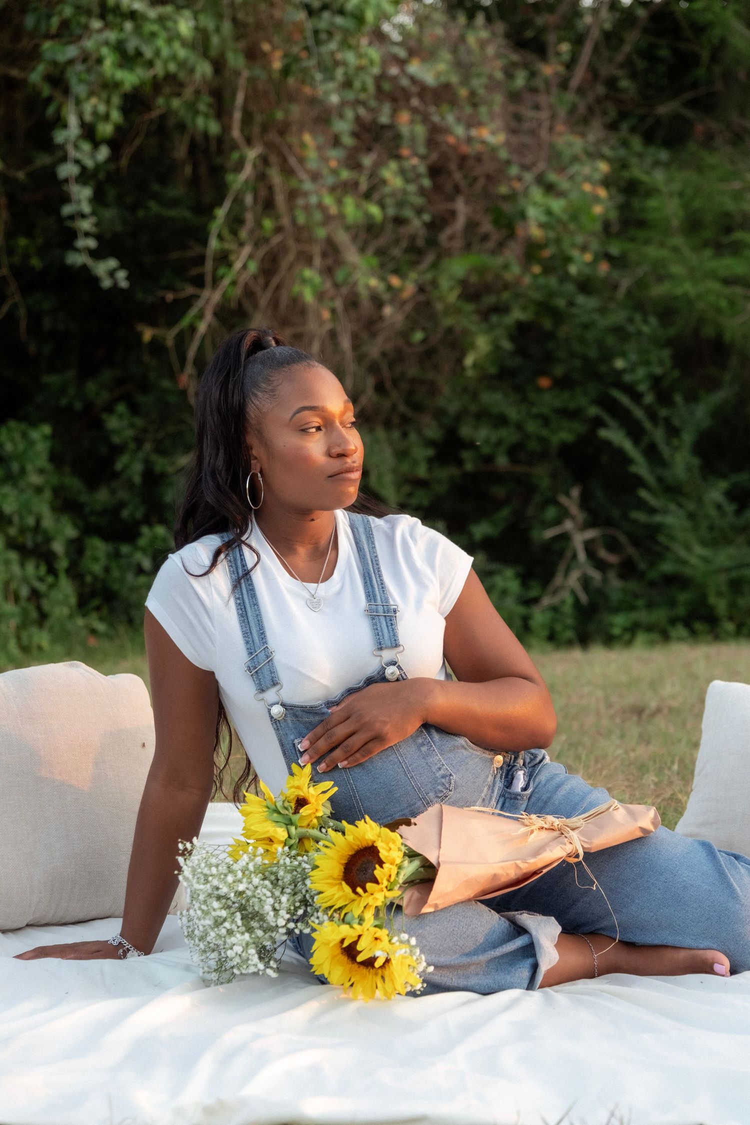 Pregnant woman in denim overalls sitting on white blanket with sunflowers, gazing away during outdoor maternity session.