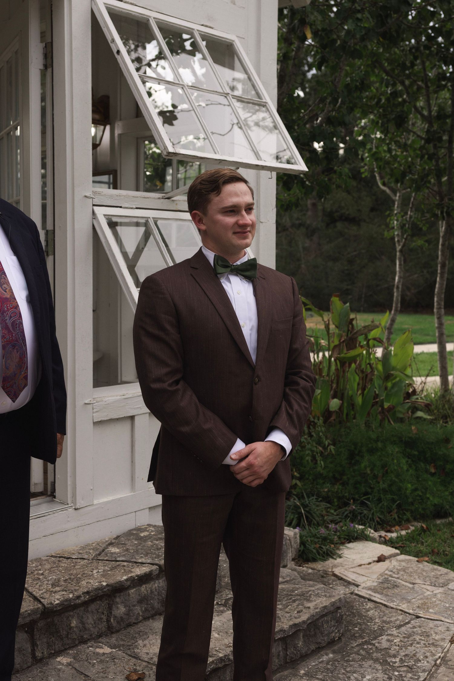 Man in brown suit with green bow tie smiling outdoors near white building with trees in background.