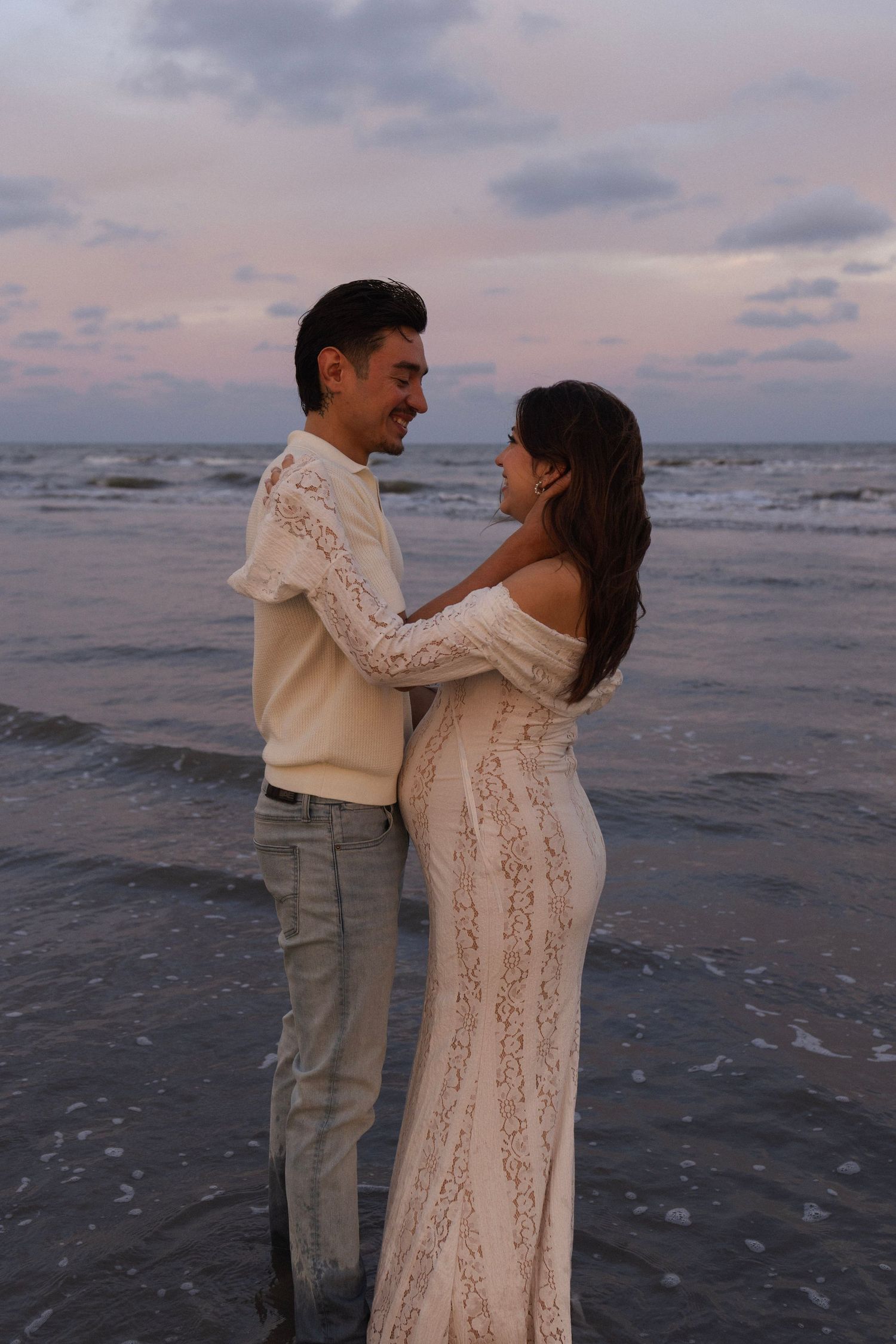 Couple embracing on a beach at sunset, woman in white lace gown, man in cream sweater, ocean waves behind them.