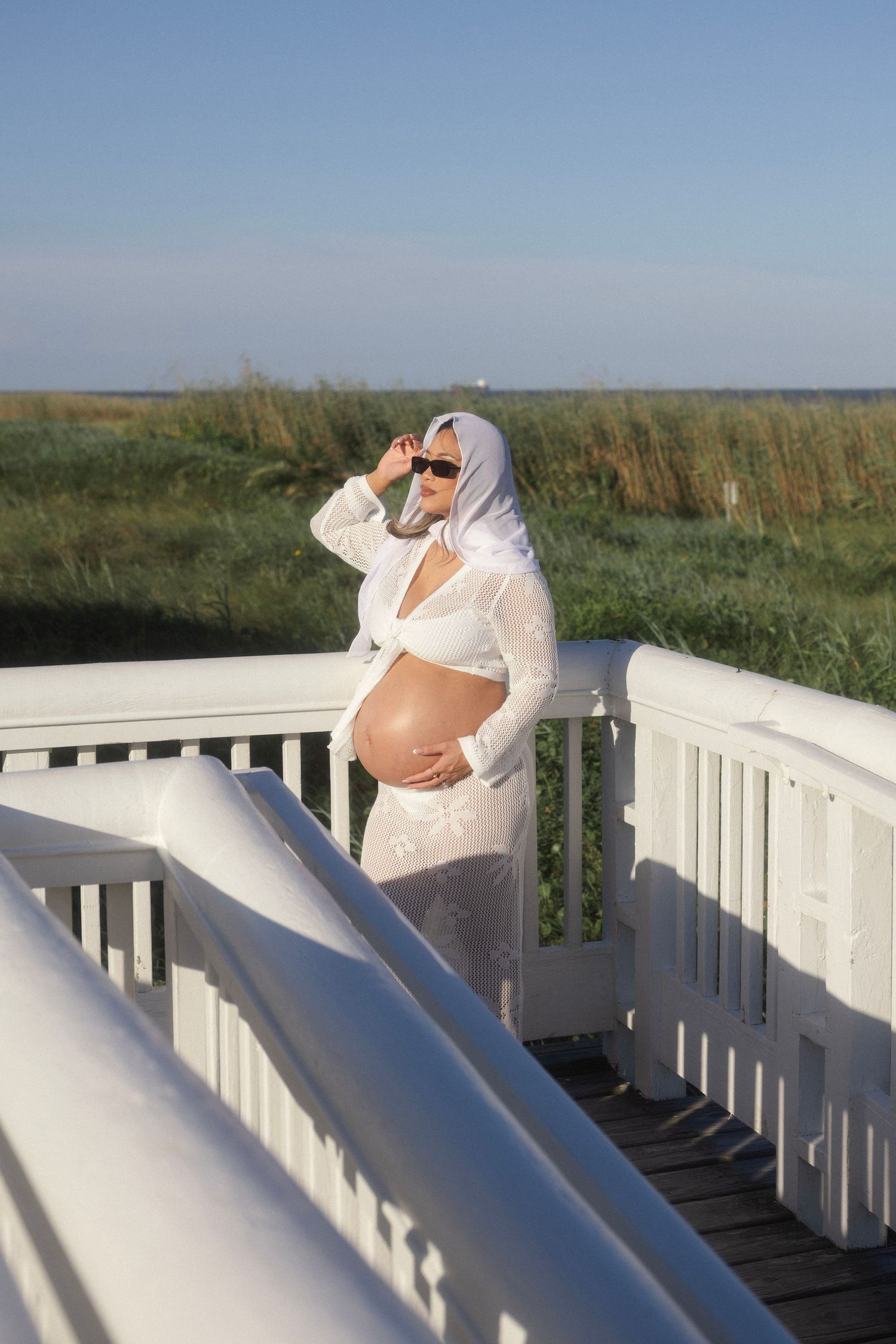 Pregnant woman in white sheer outfit and sunglasses standing on a white wooden deck overlooking green marshland.