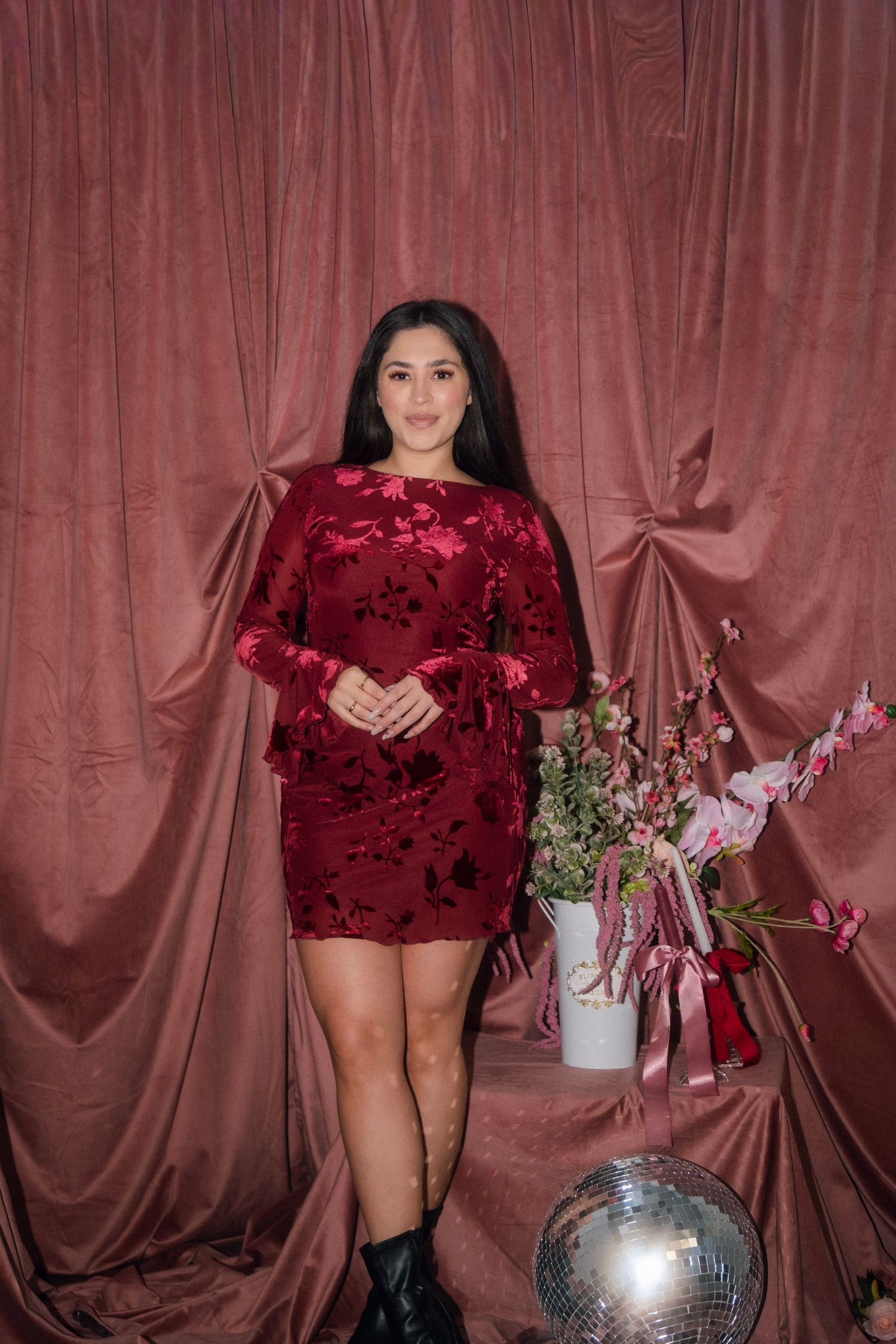 Young woman in red velvet floral mini dress poses before dusty rose curtain backdrop with flowers.