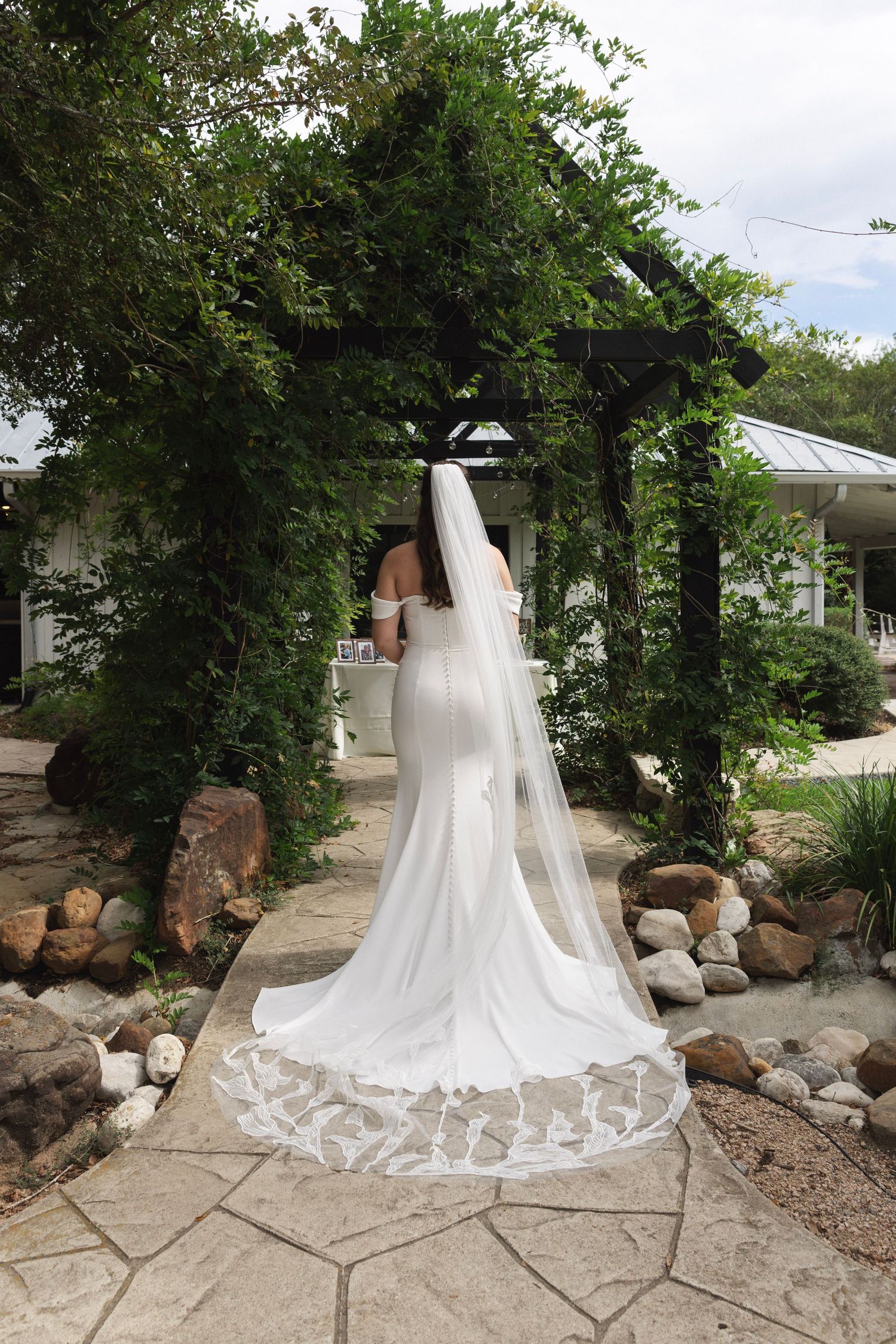 Bride in white fitted gown with long lace-trimmed veil standing under vine-covered pergola on stone path.