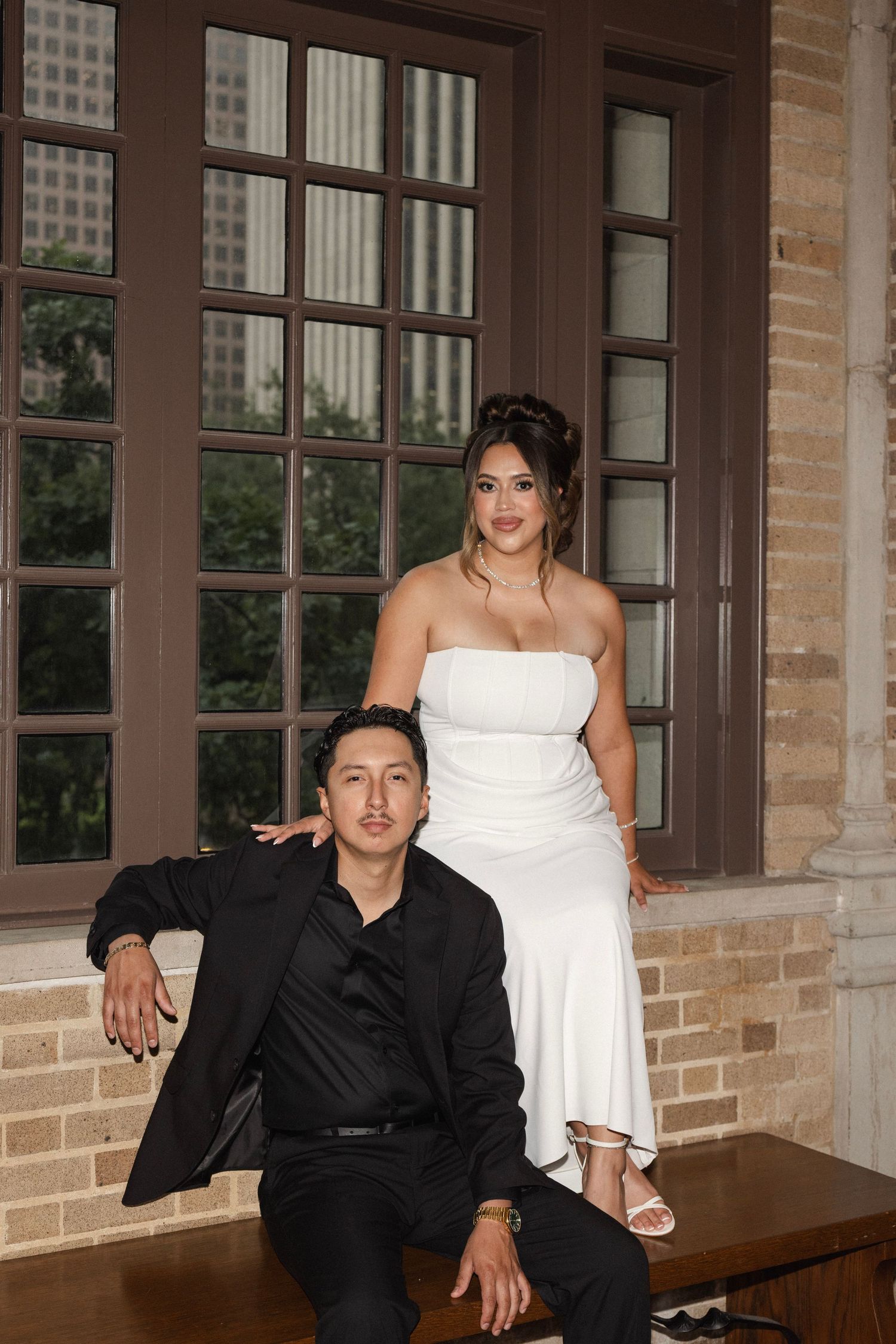 Couple posing formally, man in black suit seated, woman in white strapless gown standing behind, brick building interior.