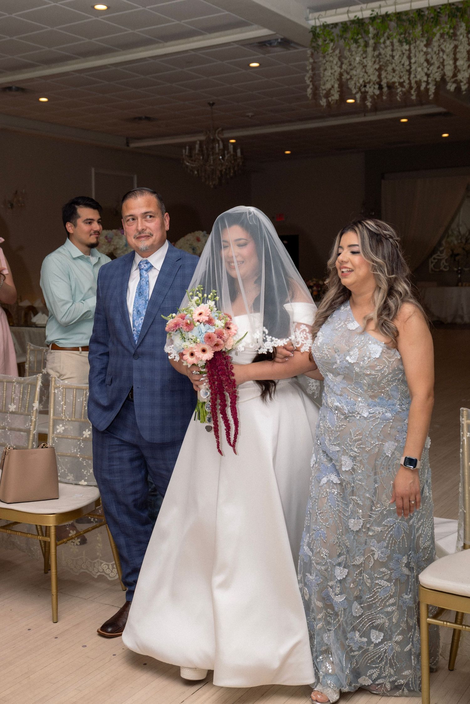 Bride walks down the aisle with parents during wedding ceremony, wearing white gown and veil holding floral bouquet.