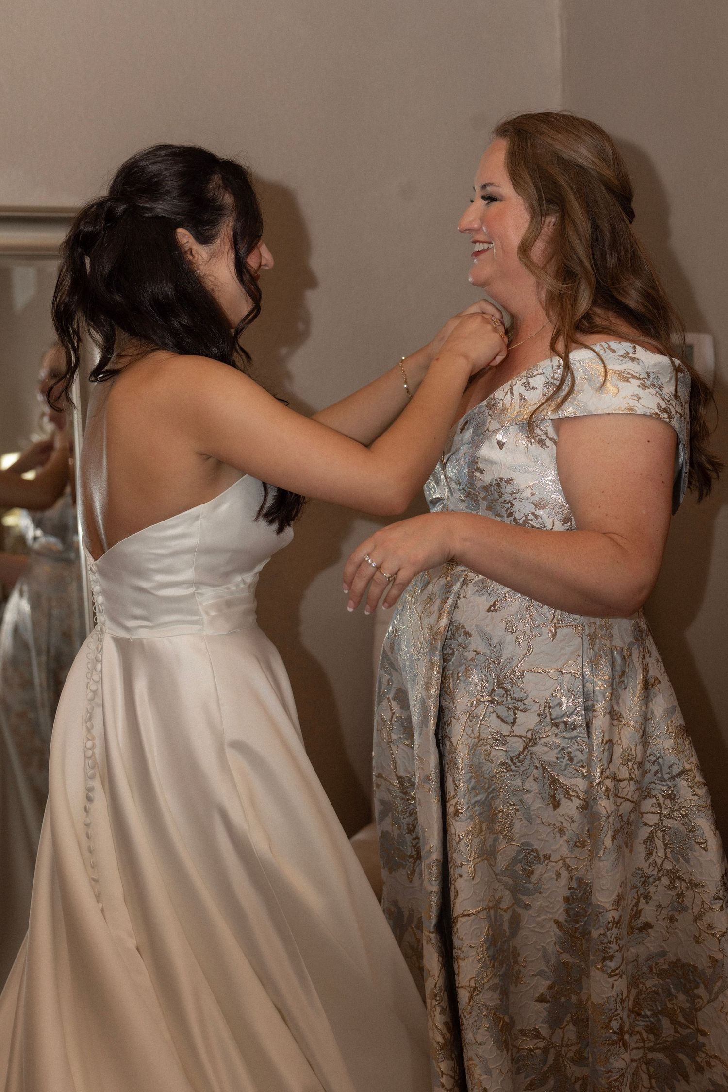 Bride in white satin gown shares a joyful moment with a woman in floral off-shoulder dress indoors.