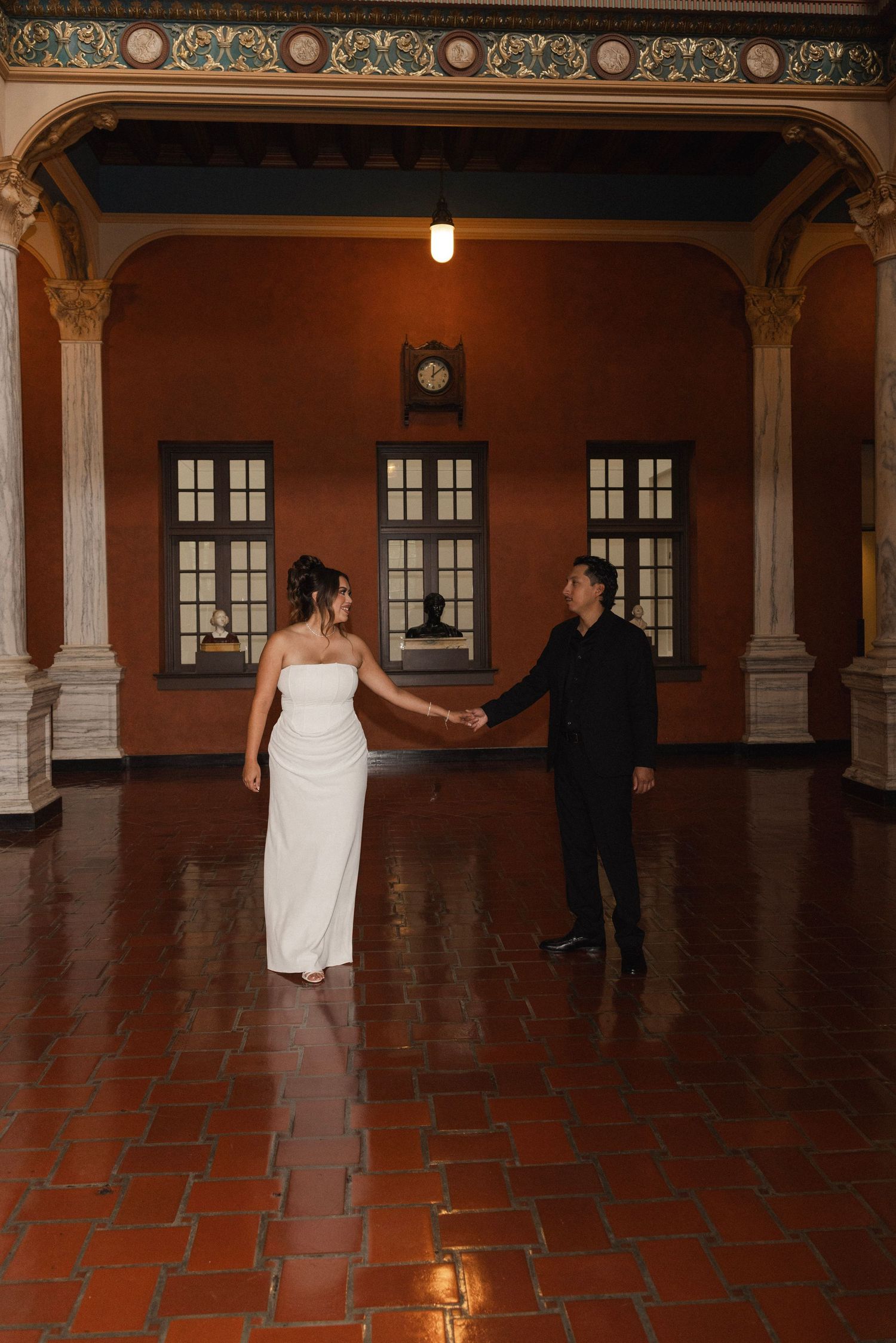 Bride in white gown and groom in black suit hold hands dancing in elegant terracotta-floored hall with arched columns.