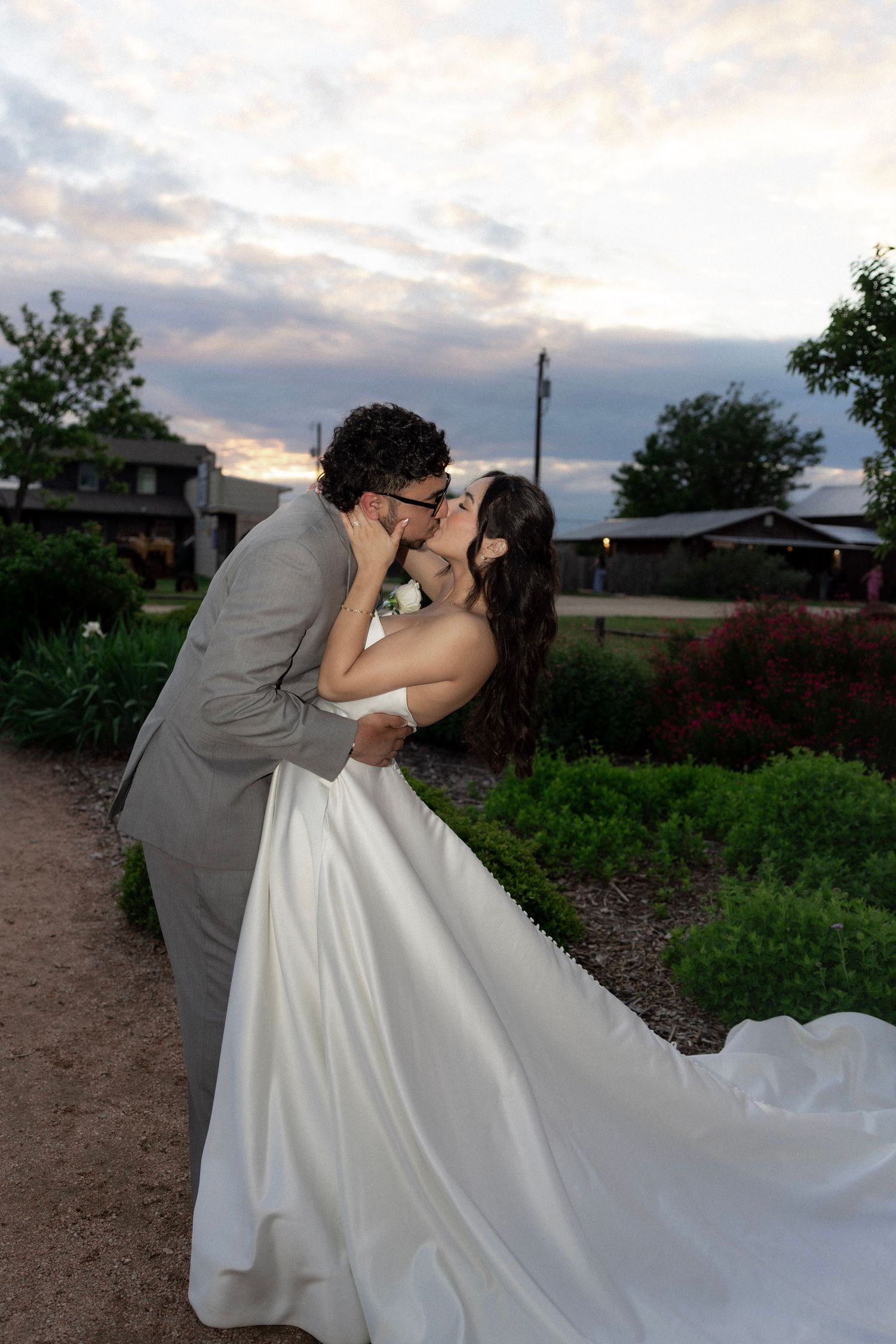 Bride and groom share a romantic dip kiss at sunset in a garden, bride in white gown, groom in gray suit.