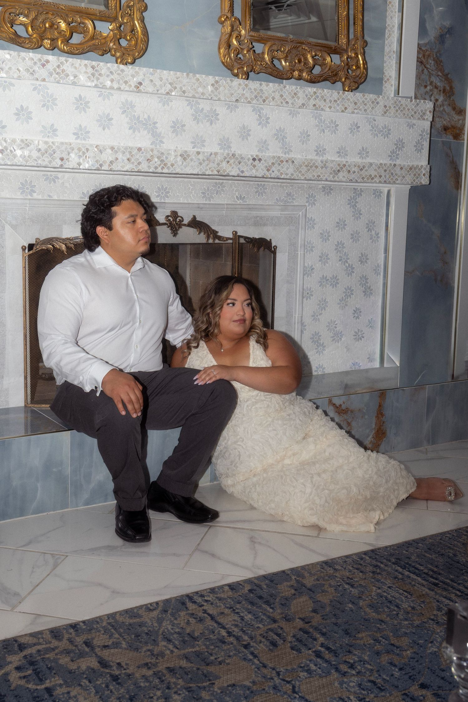 Couple in elegant attire poses together by ornate marble fireplace with gold-framed mirrors indoors.