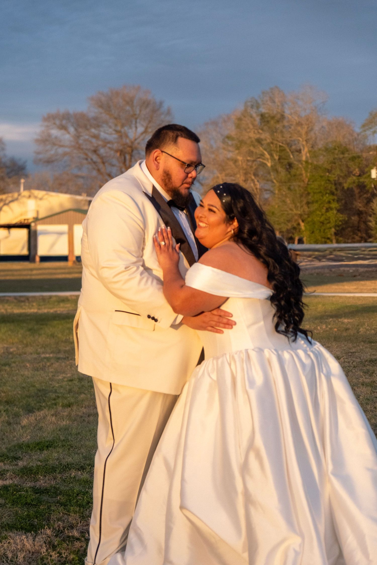 Bride and groom share romantic embrace outdoors at sunset, both dressed in white wedding attire.
