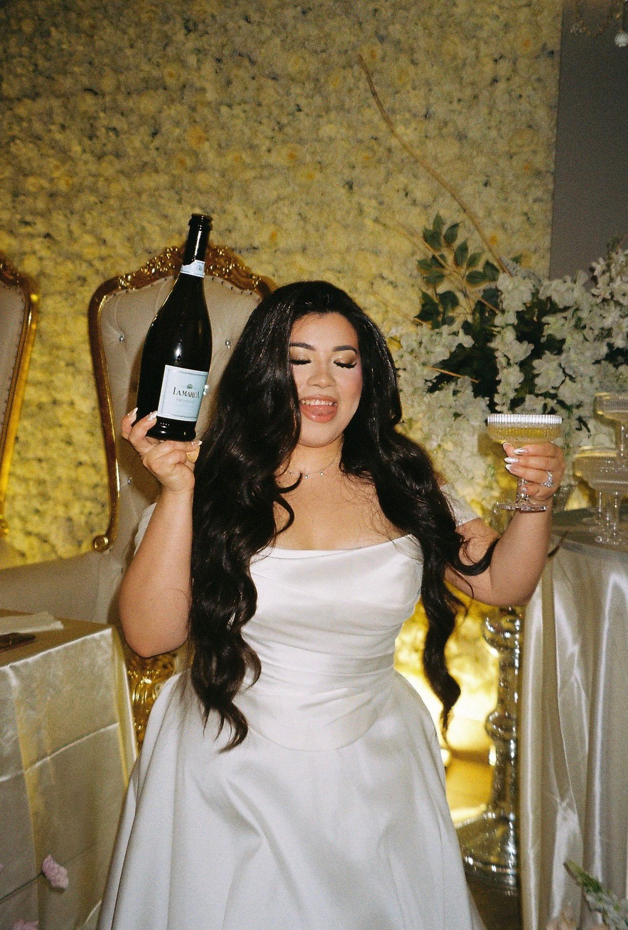 Bride in white strapless gown holds champagne bottle and glass before floral wall backdrop.