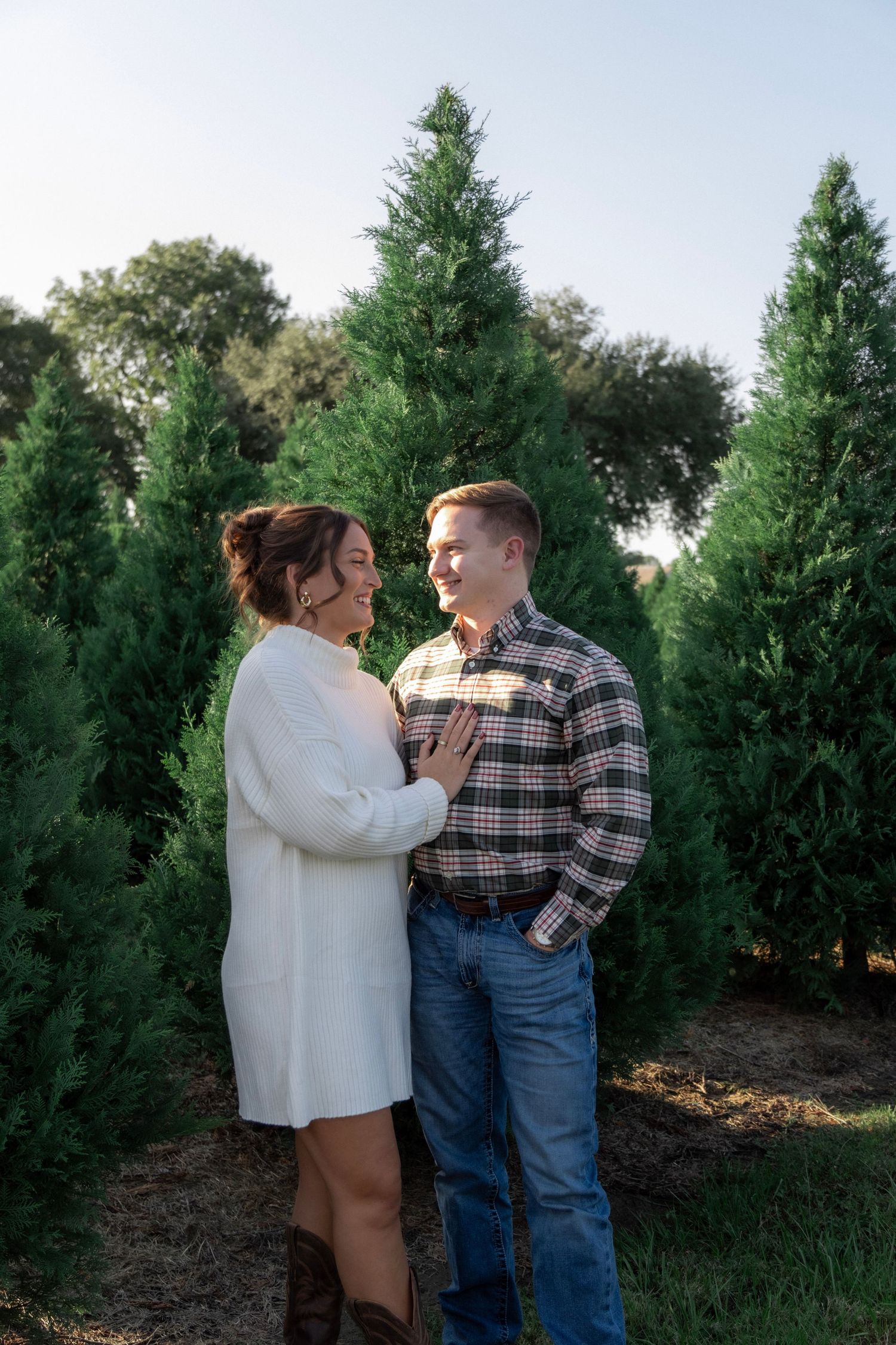 Couple smiling at each other among tall green trees at a Christmas tree farm, woman in white dress and man in plaid shirt.
