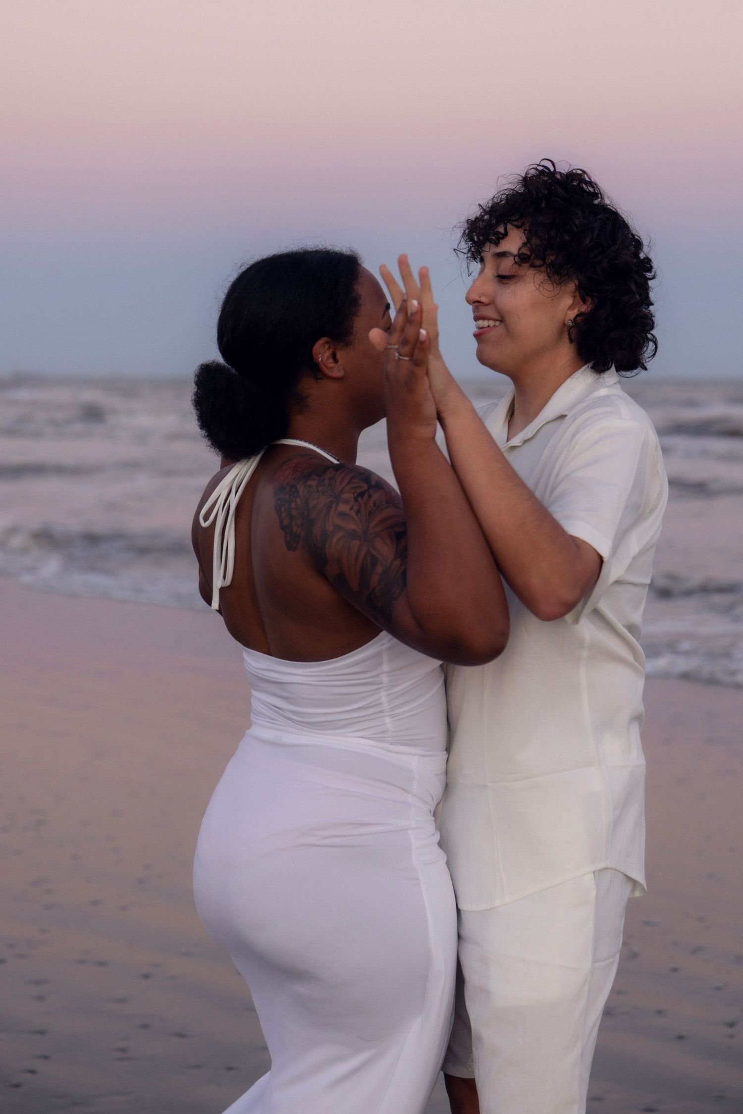 Two women in white outfits holding hands and smiling on a beach at sunset, one showing a ring.