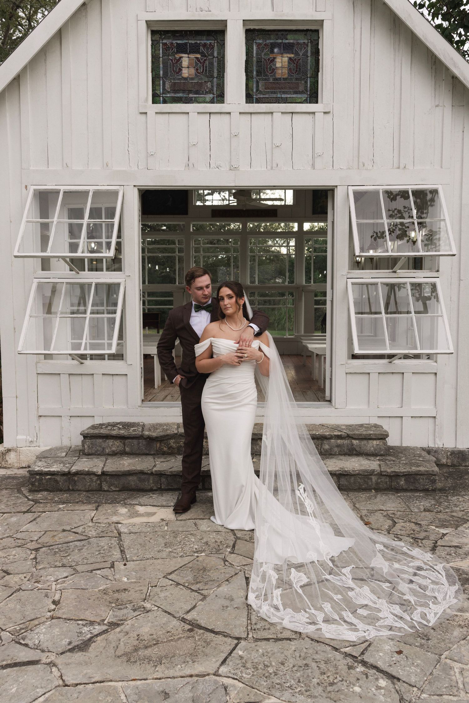 Bride in white gown with lace veil and groom in dark suit pose before a charming white chapel on stone pavers.
