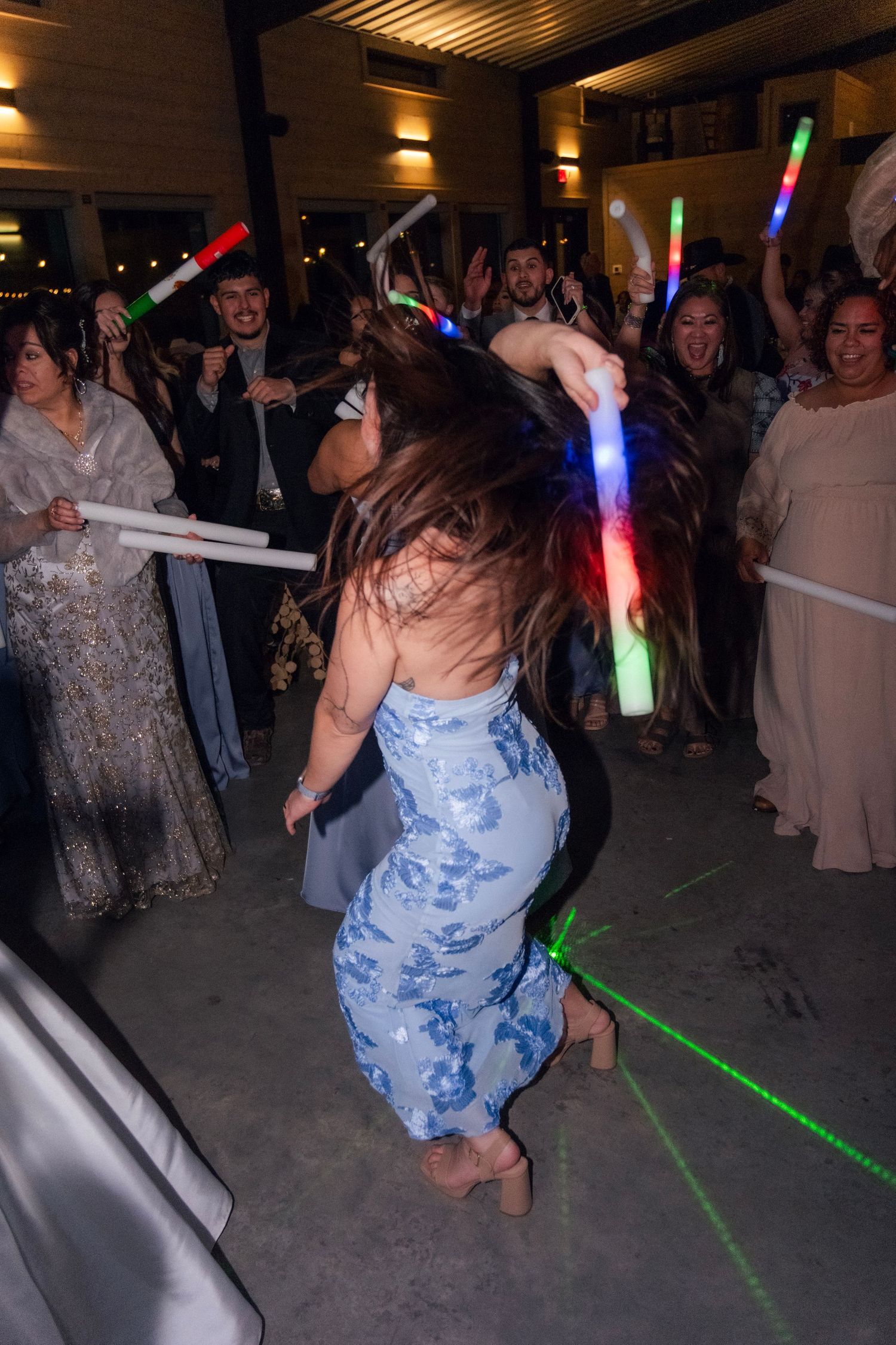 Wedding guest in blue floral dress dances with glow sticks at lively reception party.