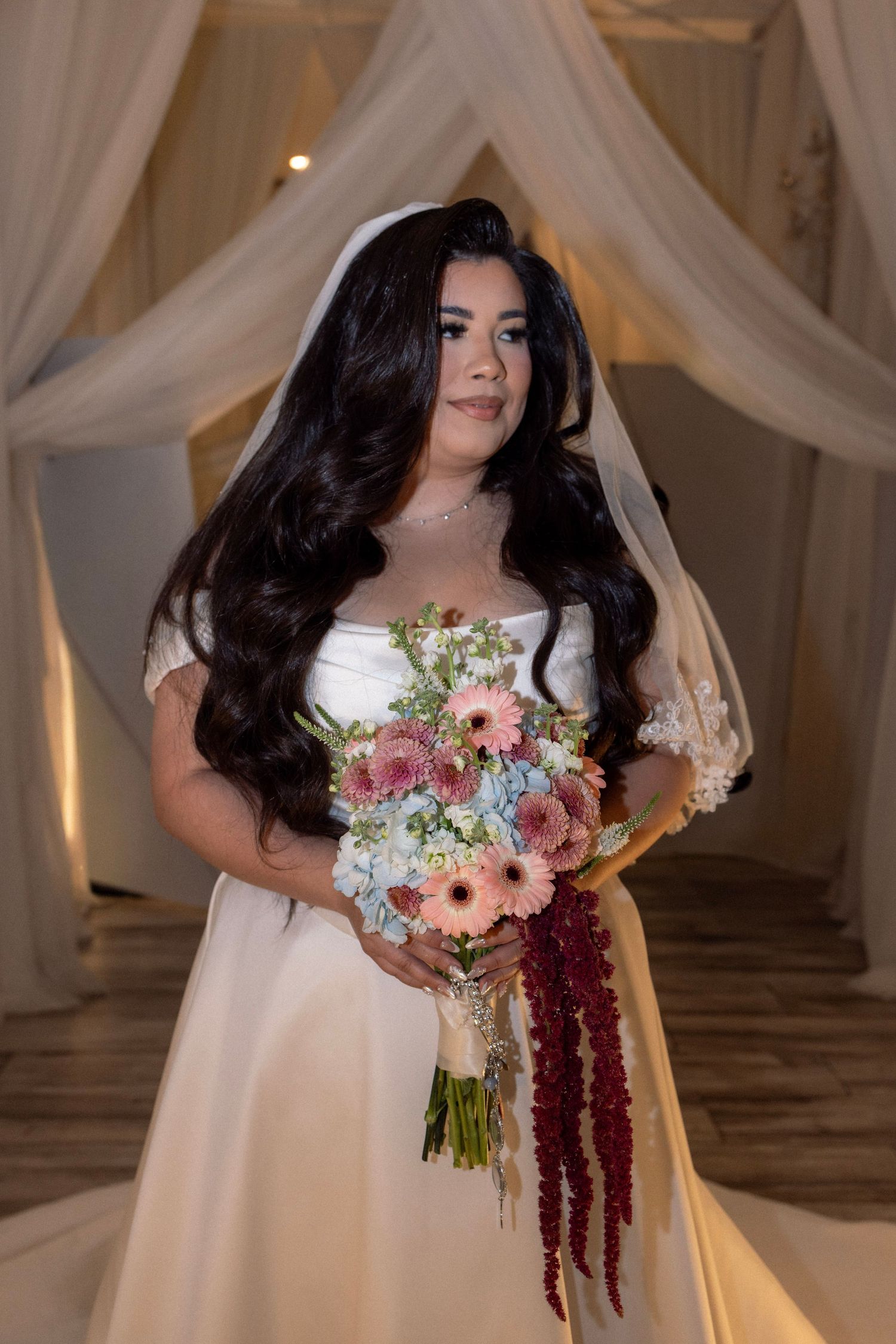 Bride in white gown holds colorful bouquet with red amaranthus under draped fabric wedding arch.