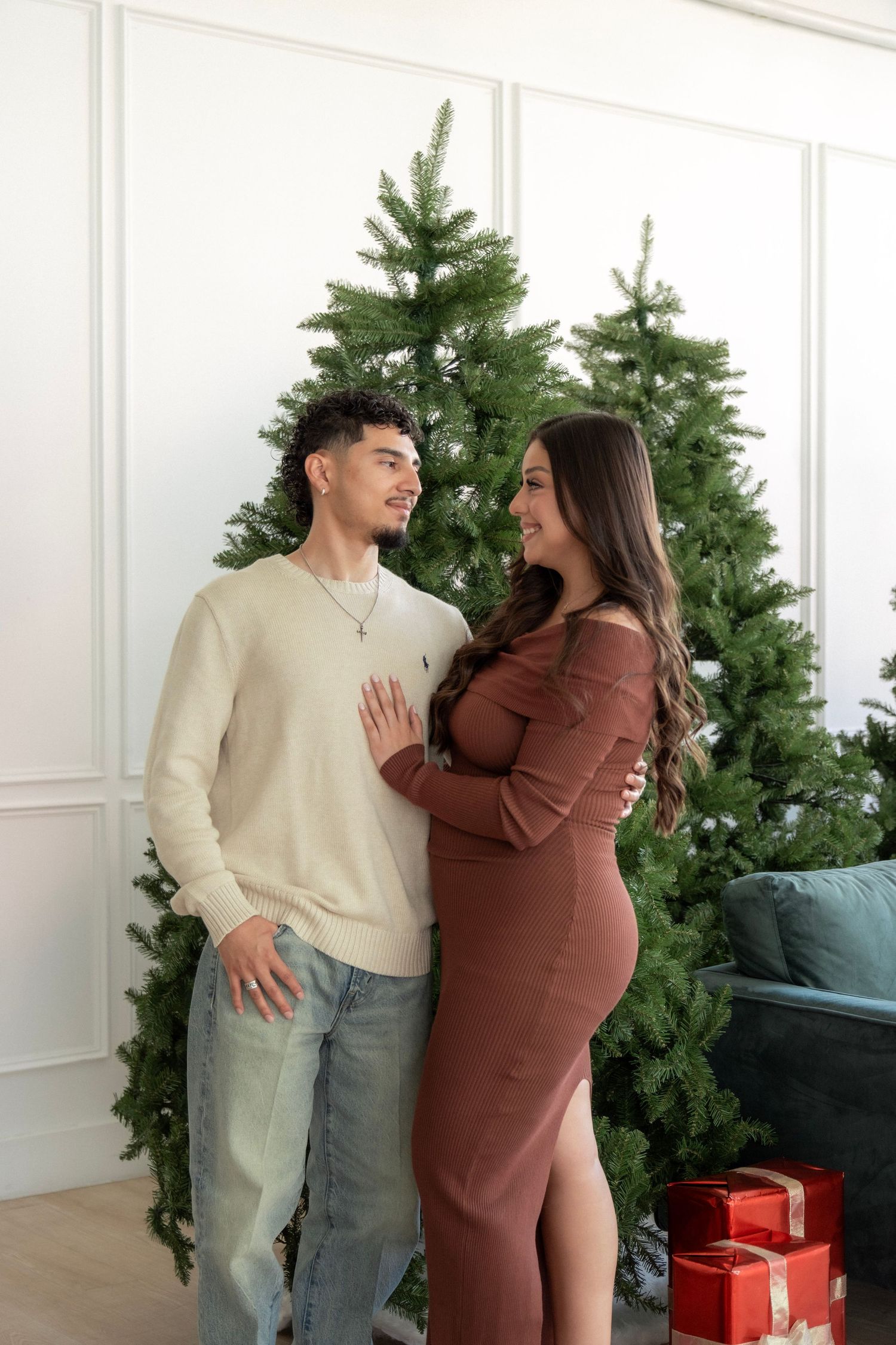 Pregnant couple poses together in front of Christmas tree with gifts, wearing cream sweater and brown dress.