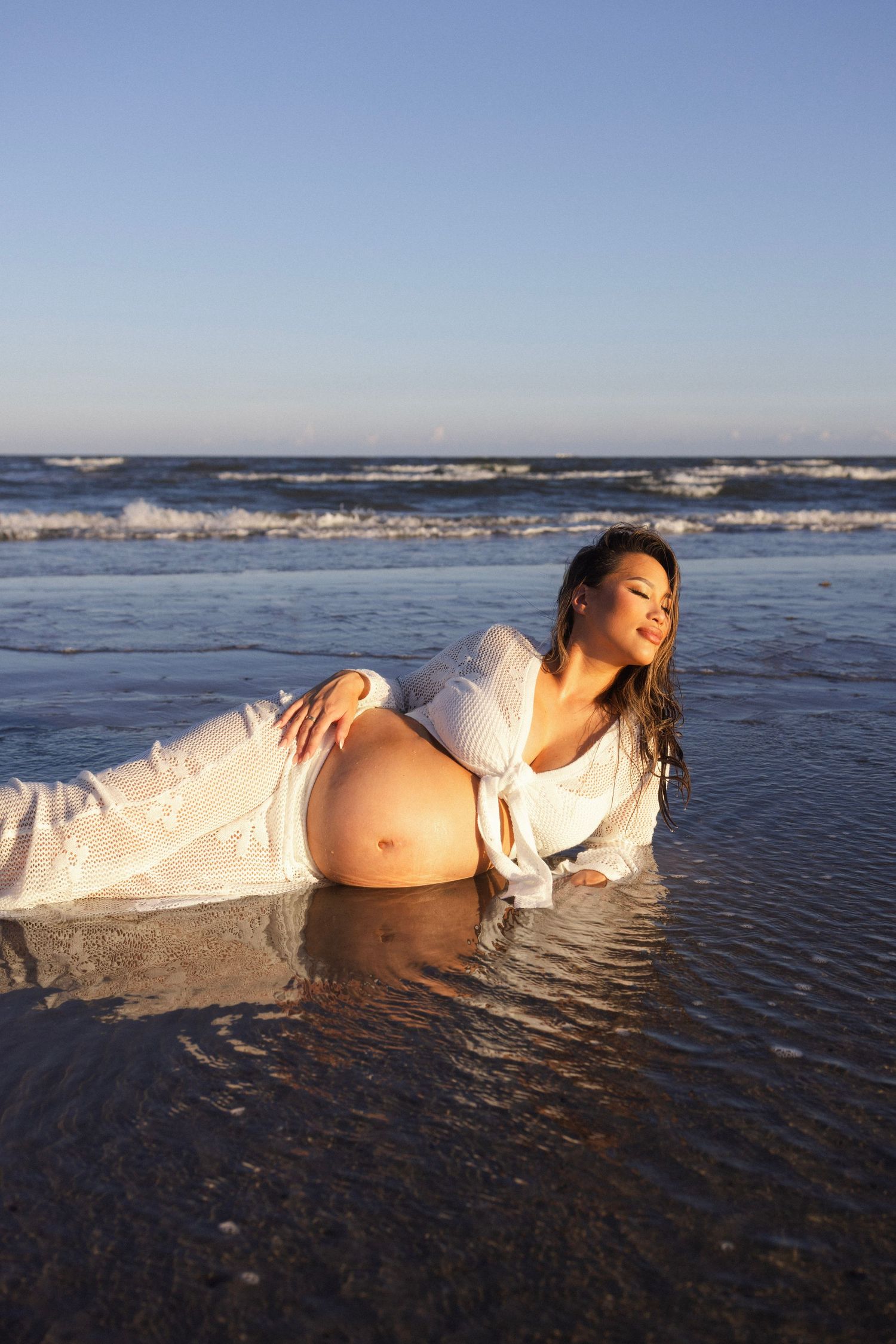 Pregnant woman in white dress lying on wet beach shore with ocean waves in background.