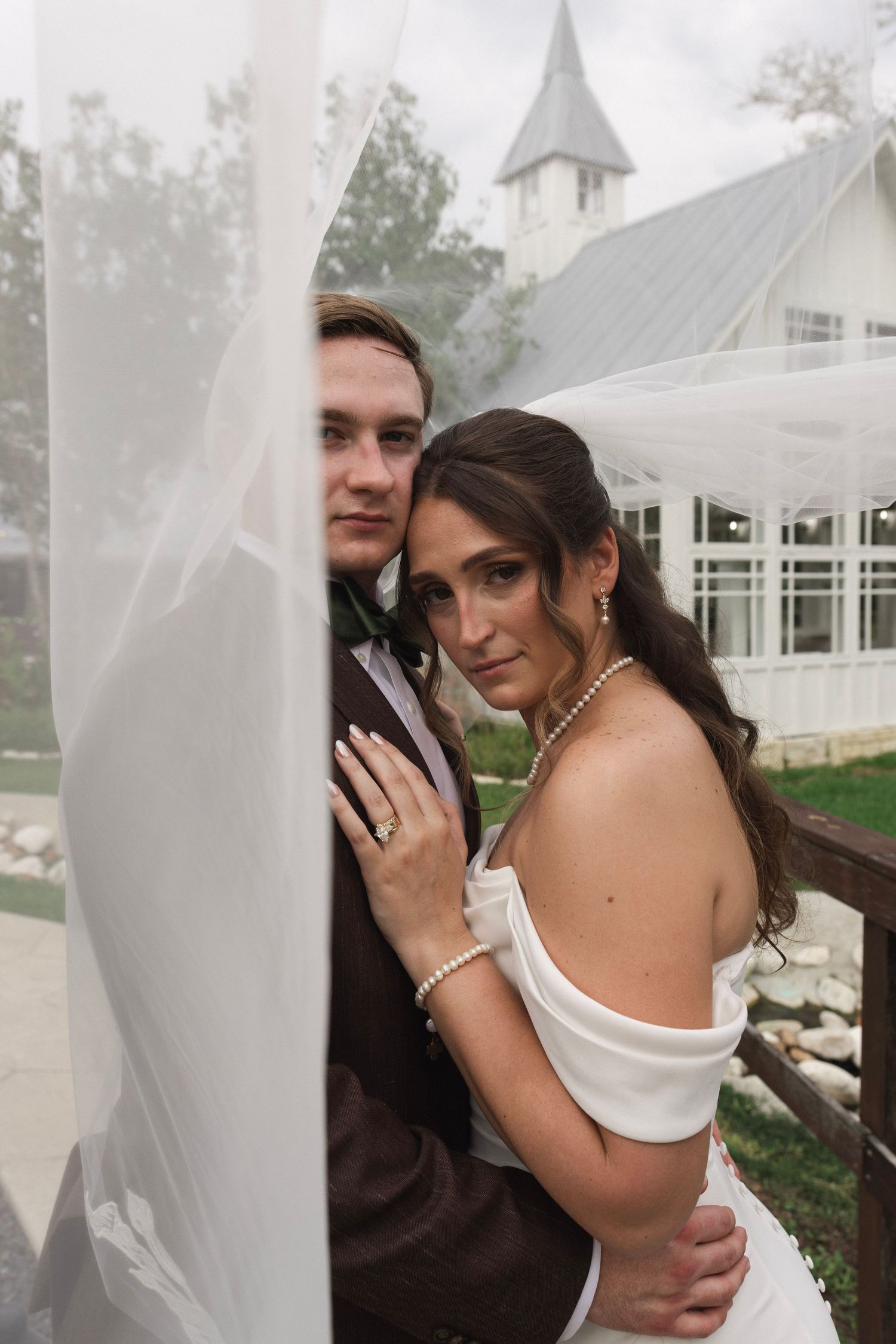 Bride and groom pose romantically under a flowing veil near a white chapel on their wedding day.