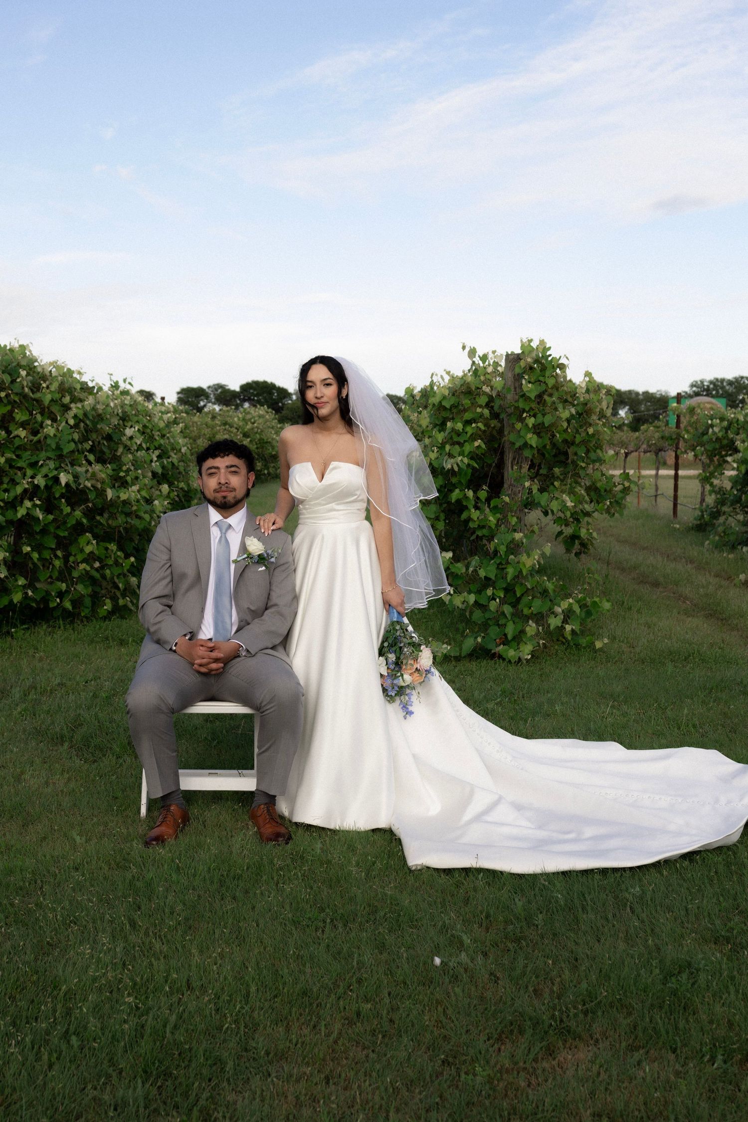 Bride holding floral bouquet stands beside seated groom in gray suit at a lush vineyard wedding.