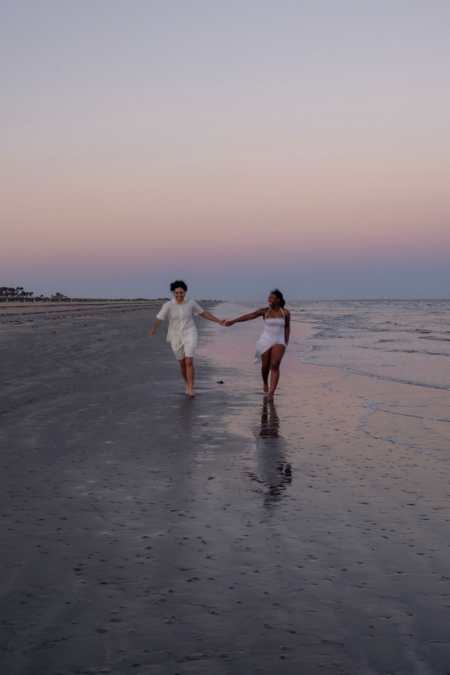Joyful couple in white outfits running hand-in-hand along a serene beach at purple sunset.