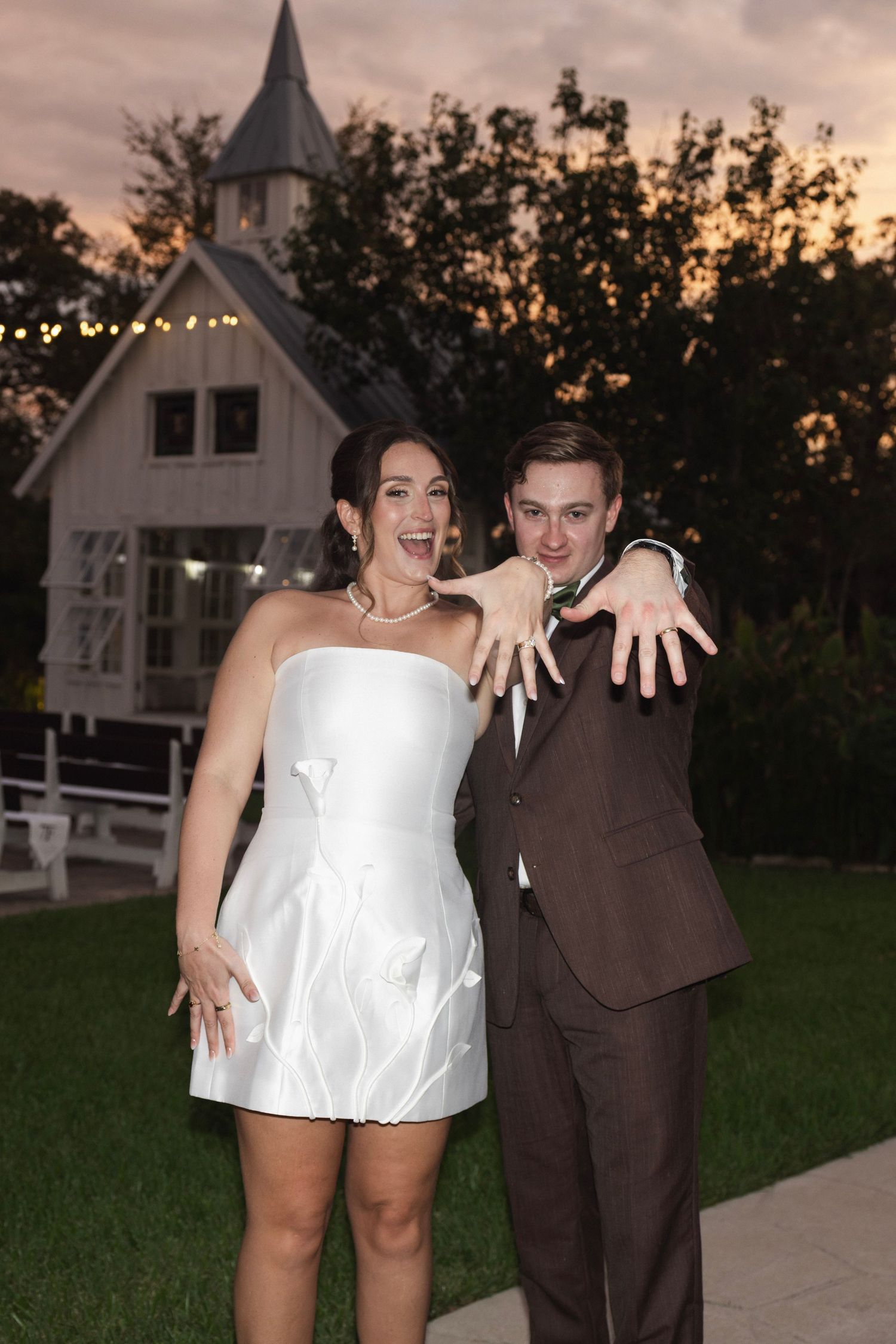 Newlyweds in white mini dress and brown suit showing off wedding rings outdoors at sunset near a white chapel.