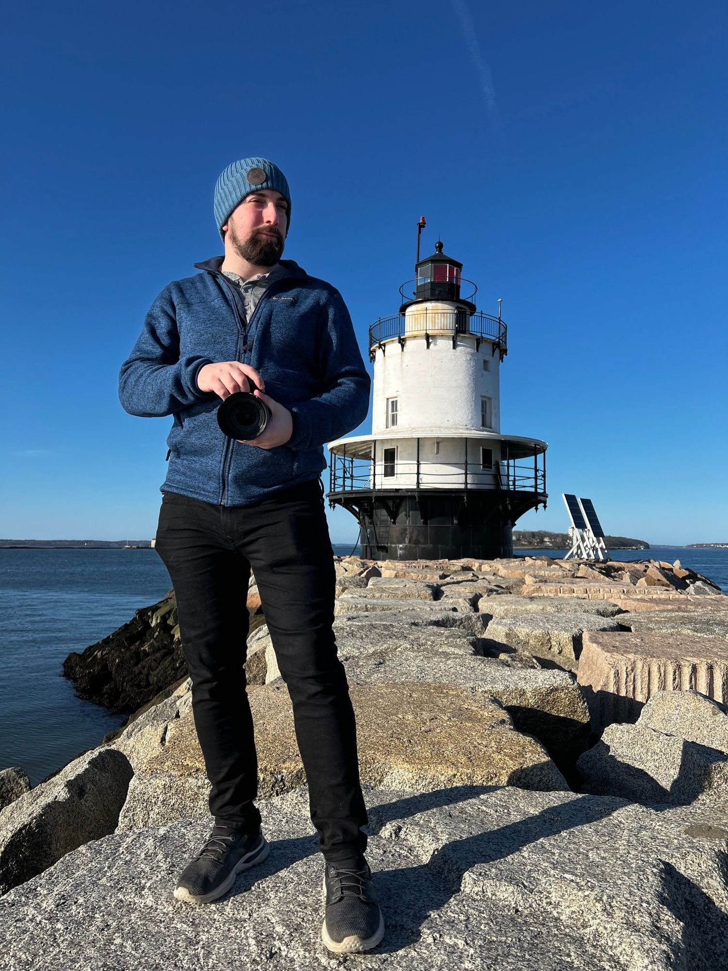 Bearded male photographer holding camera standing on rocky pier with historic lighthouse in background.