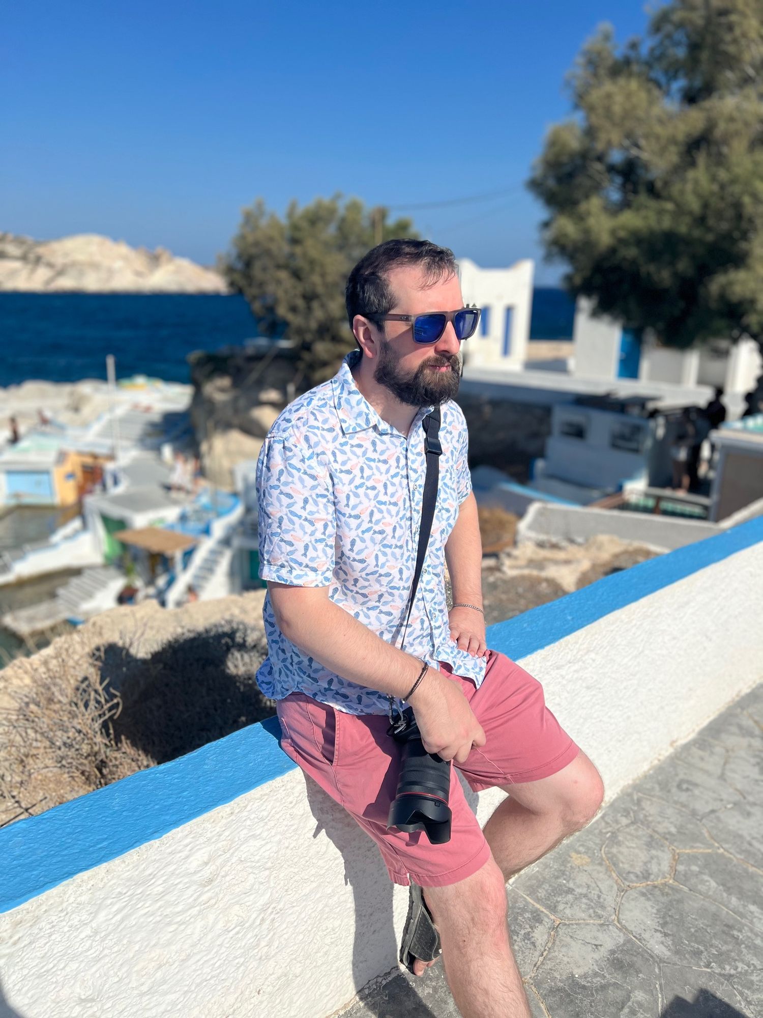 Bearded man with camera sits on blue wall overlooking Mediterranean village and sea.