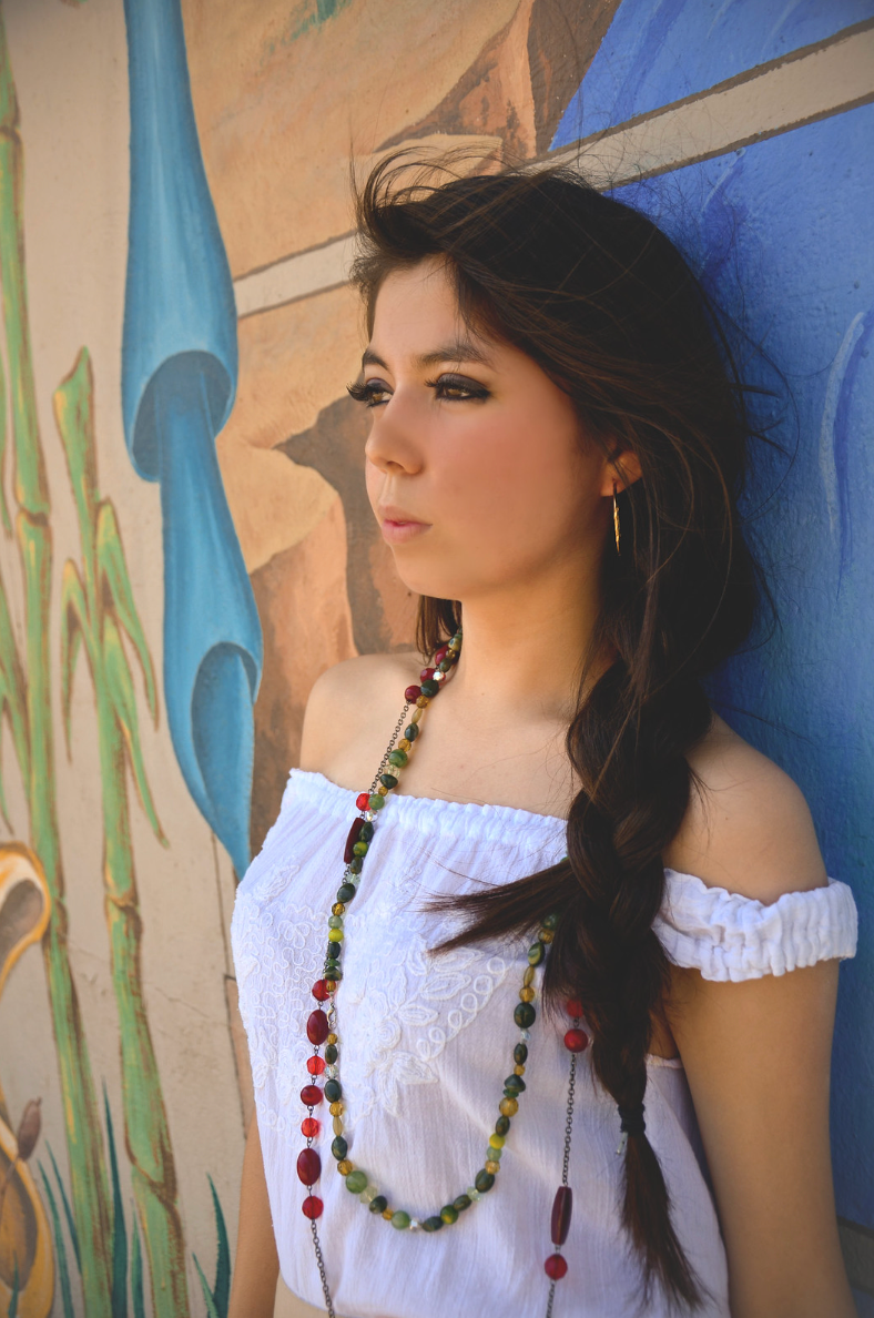 Young woman with braided hair wearing white off-shoulder top and colorful beaded necklace posing against a painted mural wall.