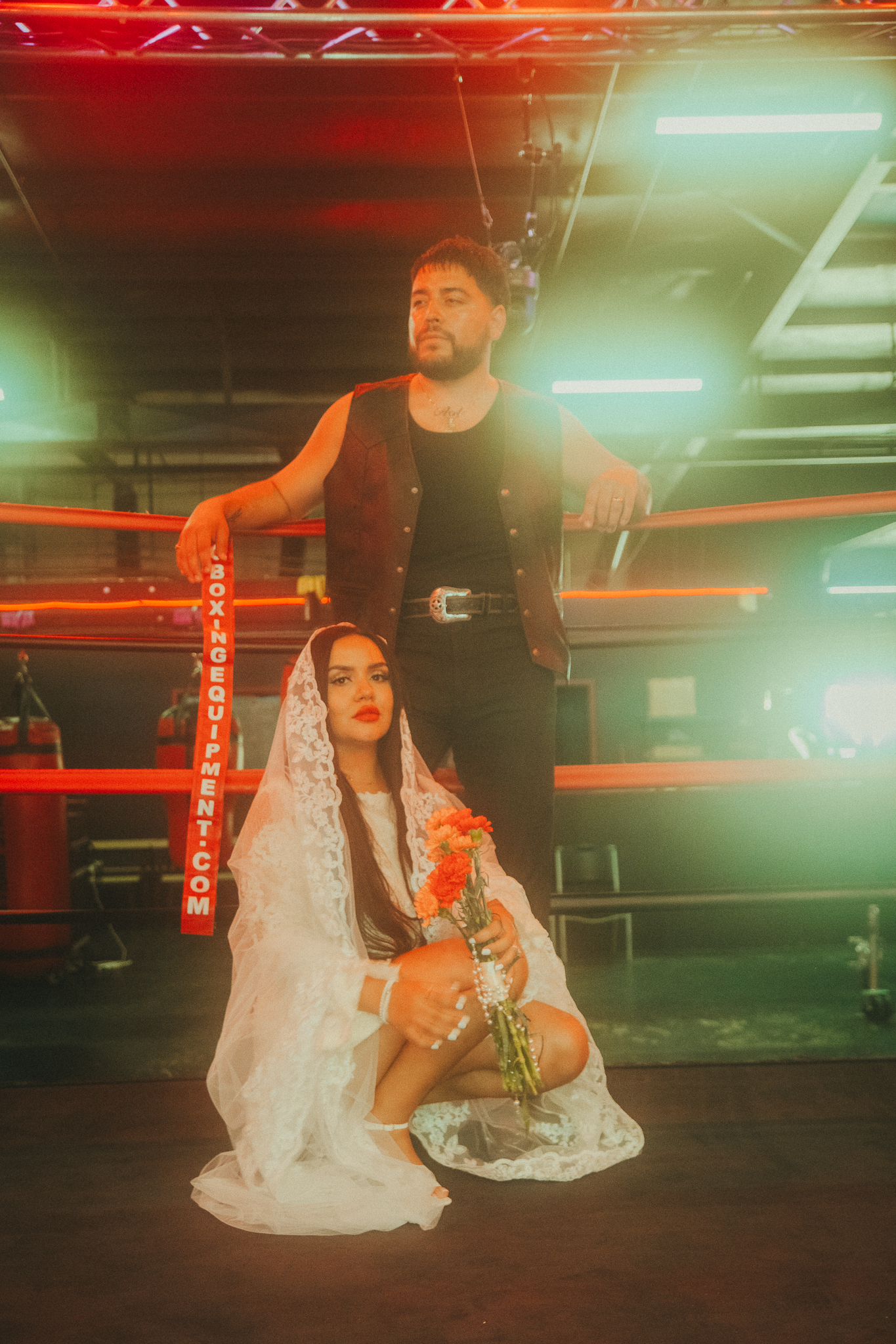 A woman in a white veil kneels holding roses beside a man in black near a boxing ring with neon lights.