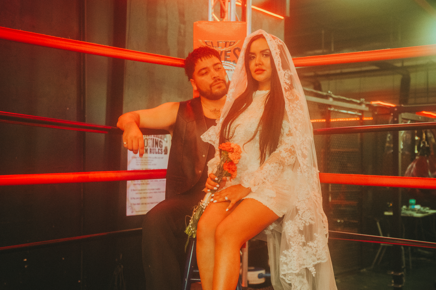 Couple posing in a boxing ring, man in black outfit and woman in white bridal gown, under red ropes.