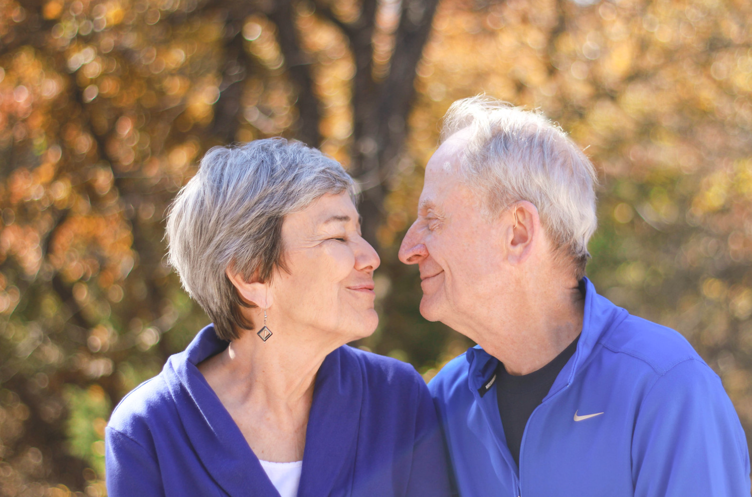 Happy senior couple in blue jackets gazing at each other with autumn trees behind them.
