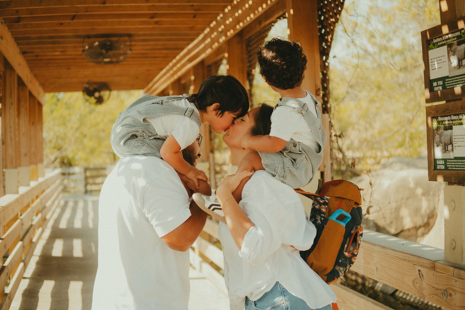 A couple shares a romantic kiss while holding a child outdoors on a sunny wooden deck.