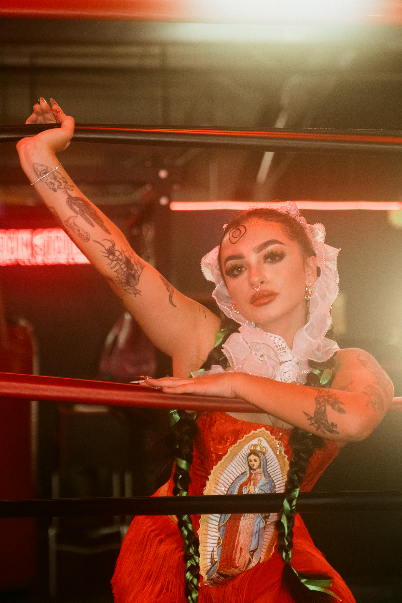 Tattooed woman in red dress with Virgin Mary print poses by wrestling ring ropes under neon lights.
